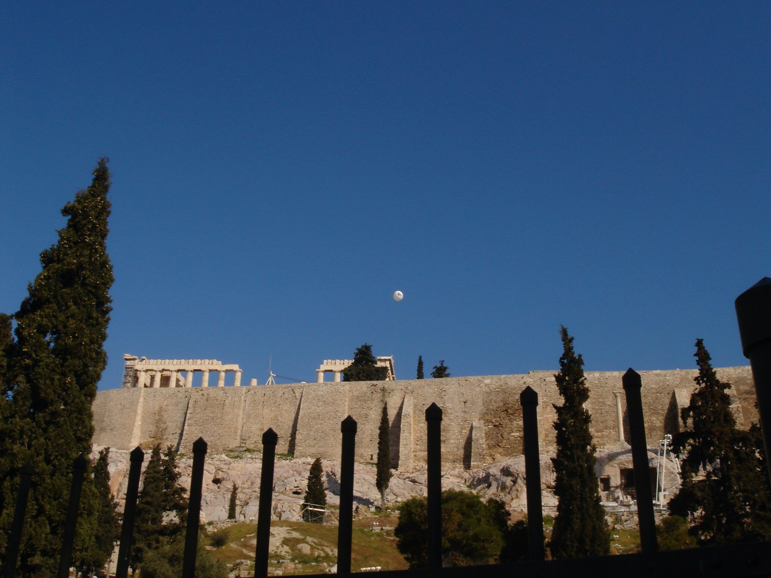 The Parthenon seen from below the Acropolis walls, with tall dark cypress trees and a fence in the foreground.