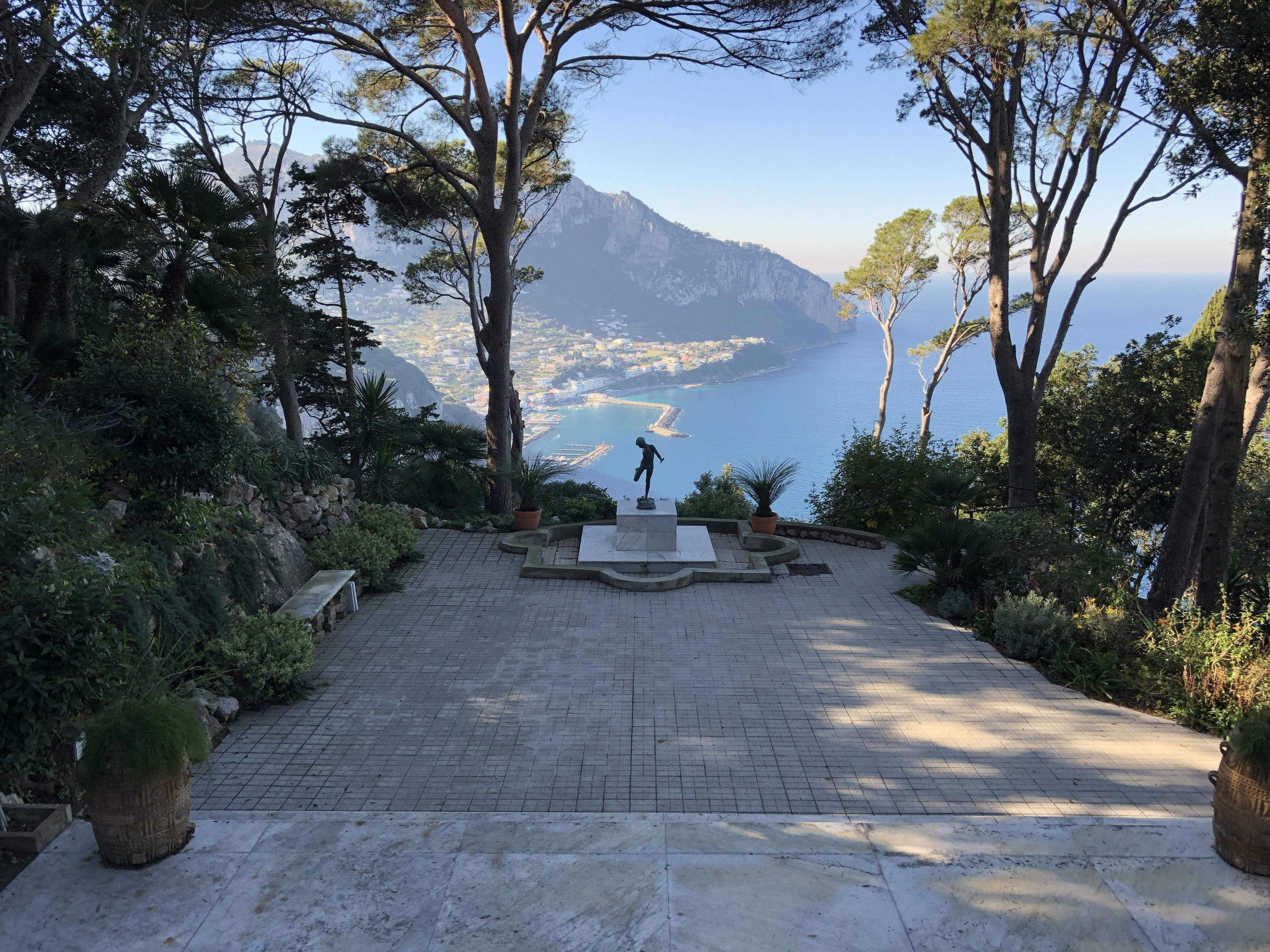 Statue on a terrace overlooking the Bay of Naples, with pine trees framing the view of Capri's cliffs and Marina Grande below.