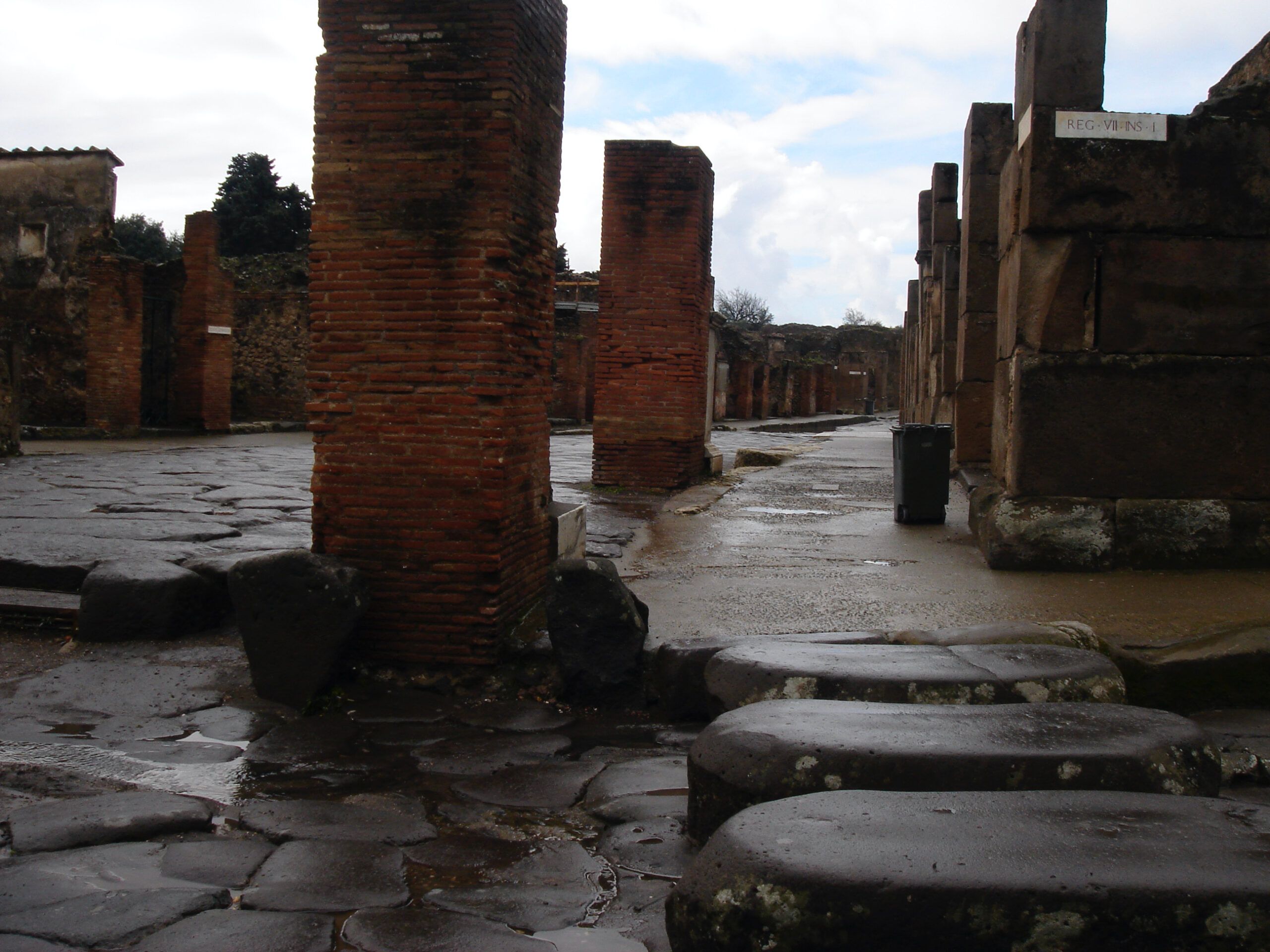 Wet stone-paved street in Pompeii lined with brick columns and stepping stones, under a patchy sky with visible street signage.