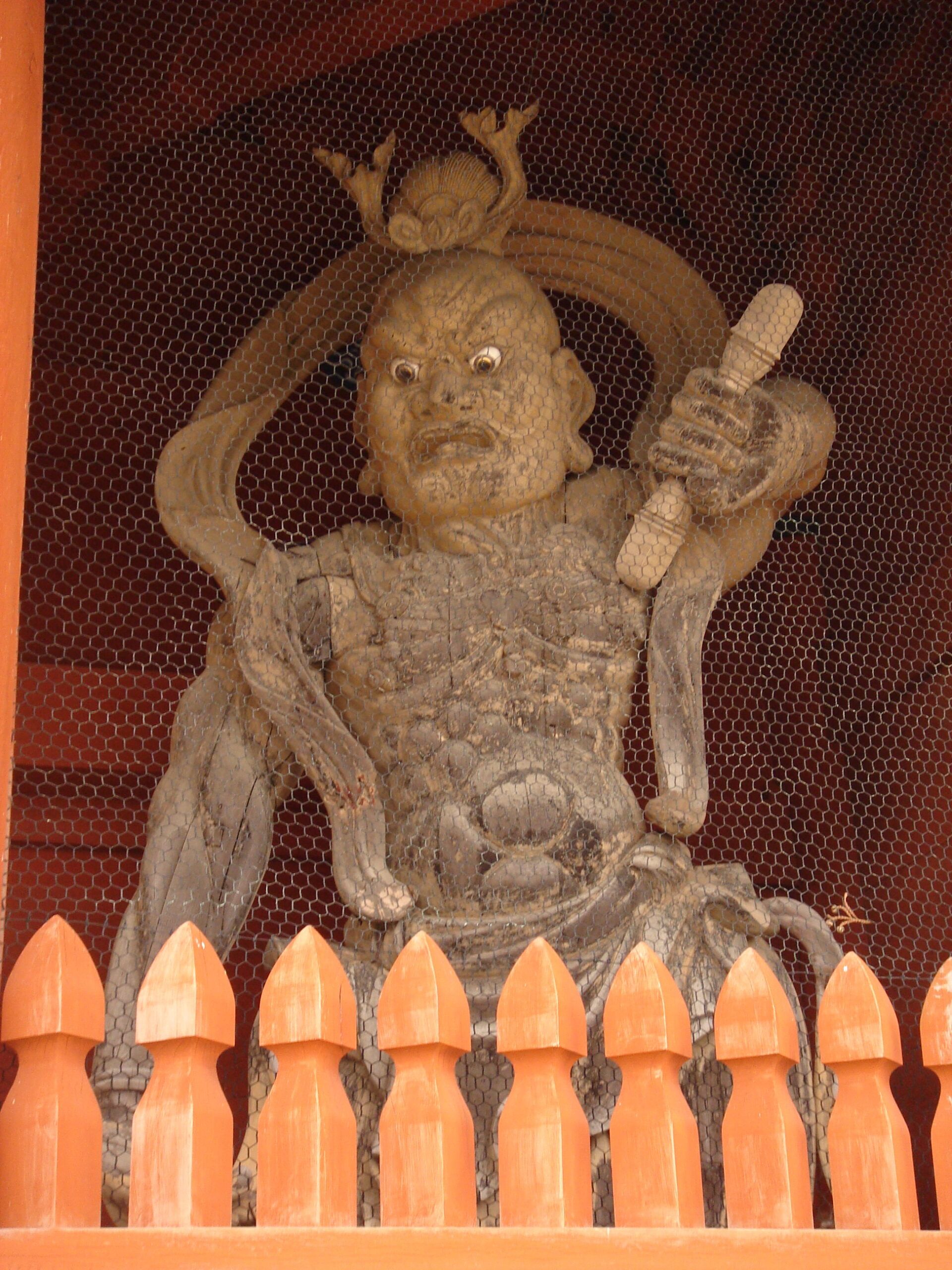 A guardian statue behind mesh and a red fence, holding a weapon and baring its teeth.