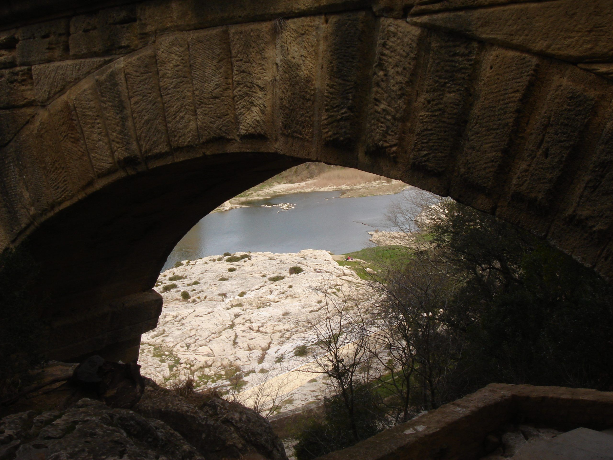 View of the Gardon River and rocky riverbank seen through the underside of a stone arch at the Pont du Gard.