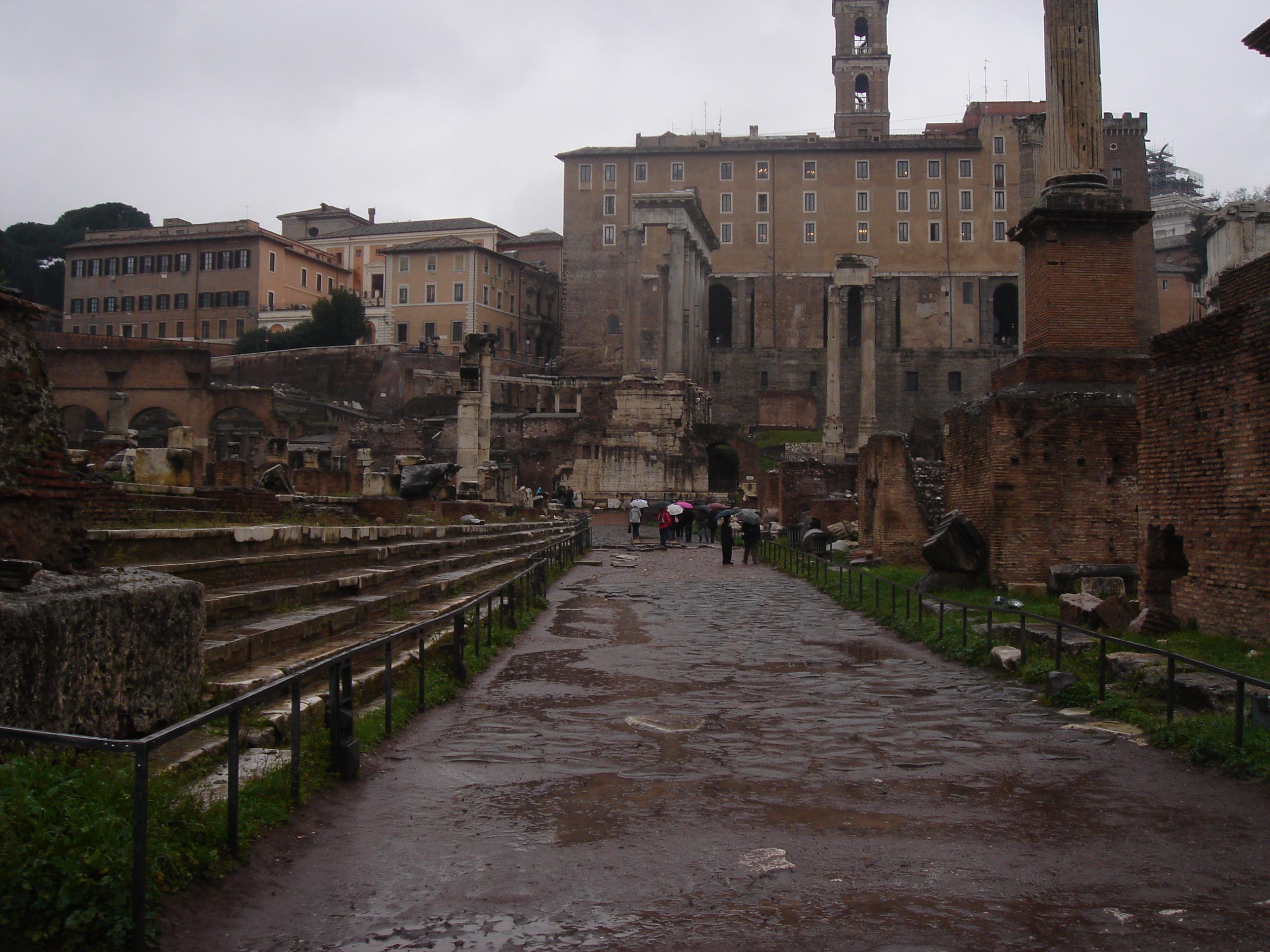 A wide view of the Roman Forum on a rainy day, with ancient ruins, scattered columns, and tourists walking along the Via Sacra toward the Curia.