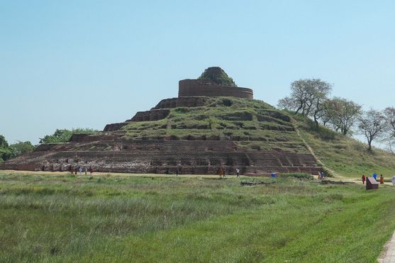 View of the massive brick stupa at Kesariya, overgrown with grass and visited by small groups of tourists on a sunny day.