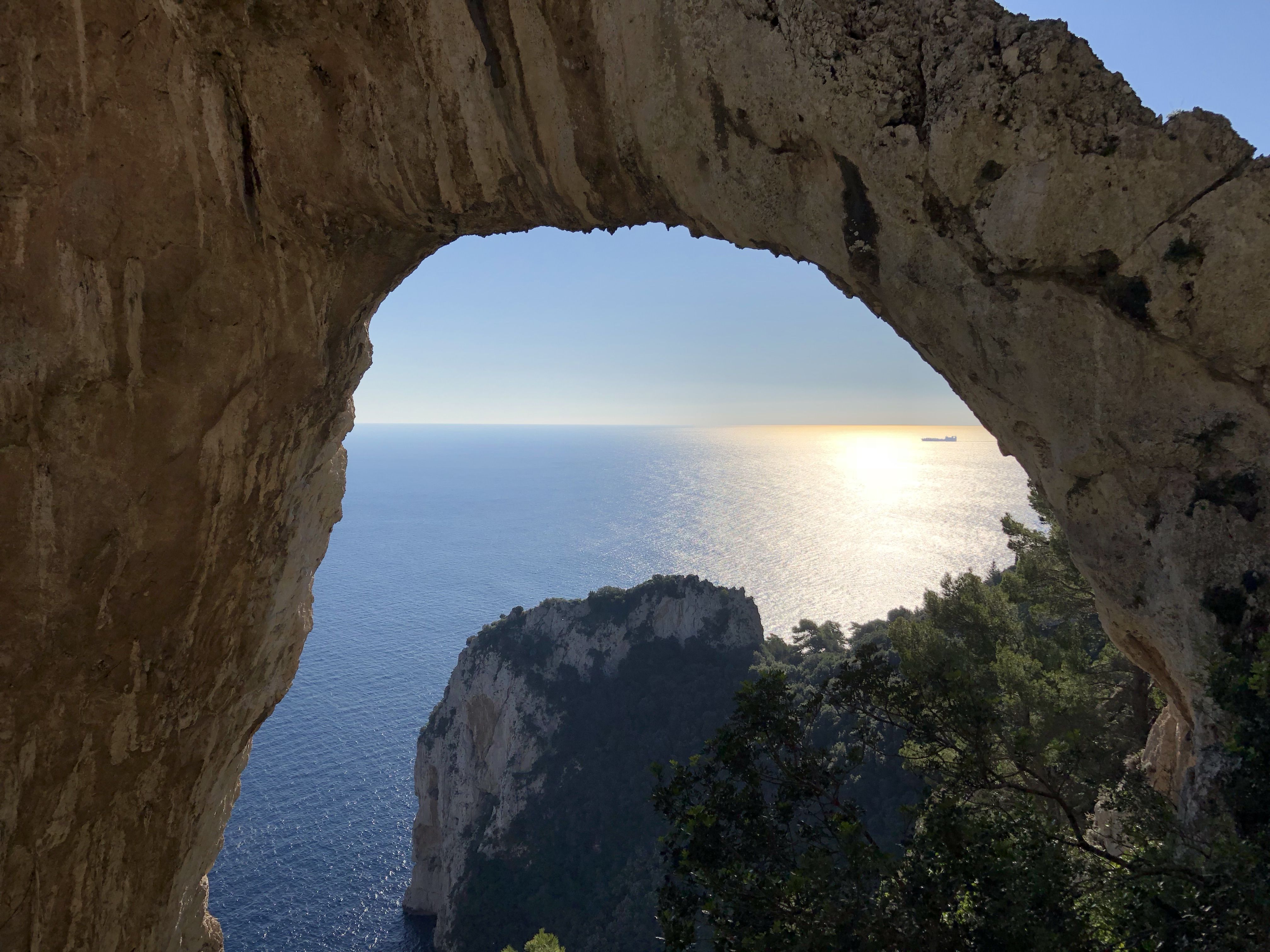 Sunlit sea seen through the rocky stone arch of the Arco Naturale on Capri, with a cargo ship visible on the horizon.