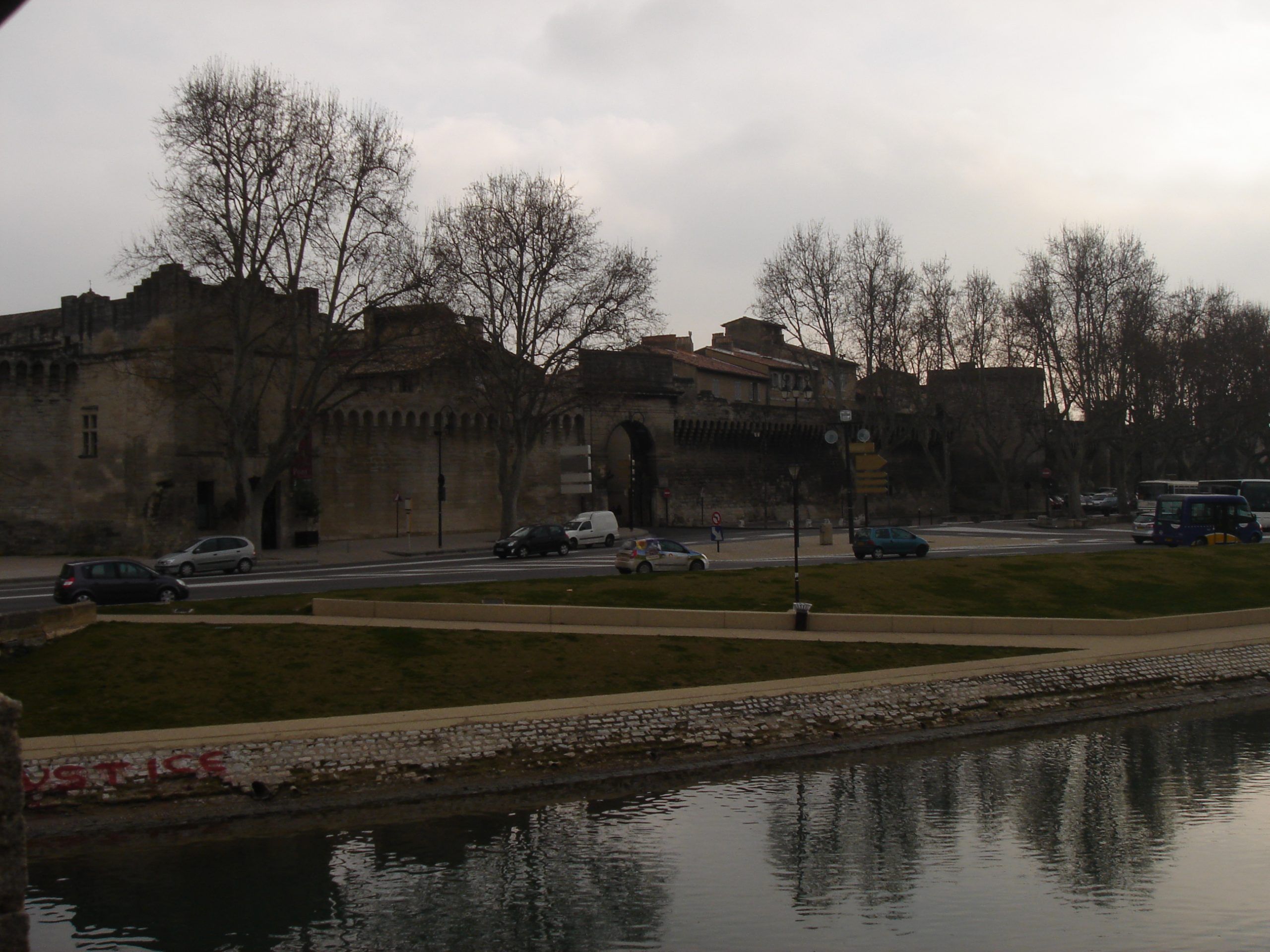 View of the medieval city walls of Avignon, seen from across the Rhône River with cars and buses passing along the road.