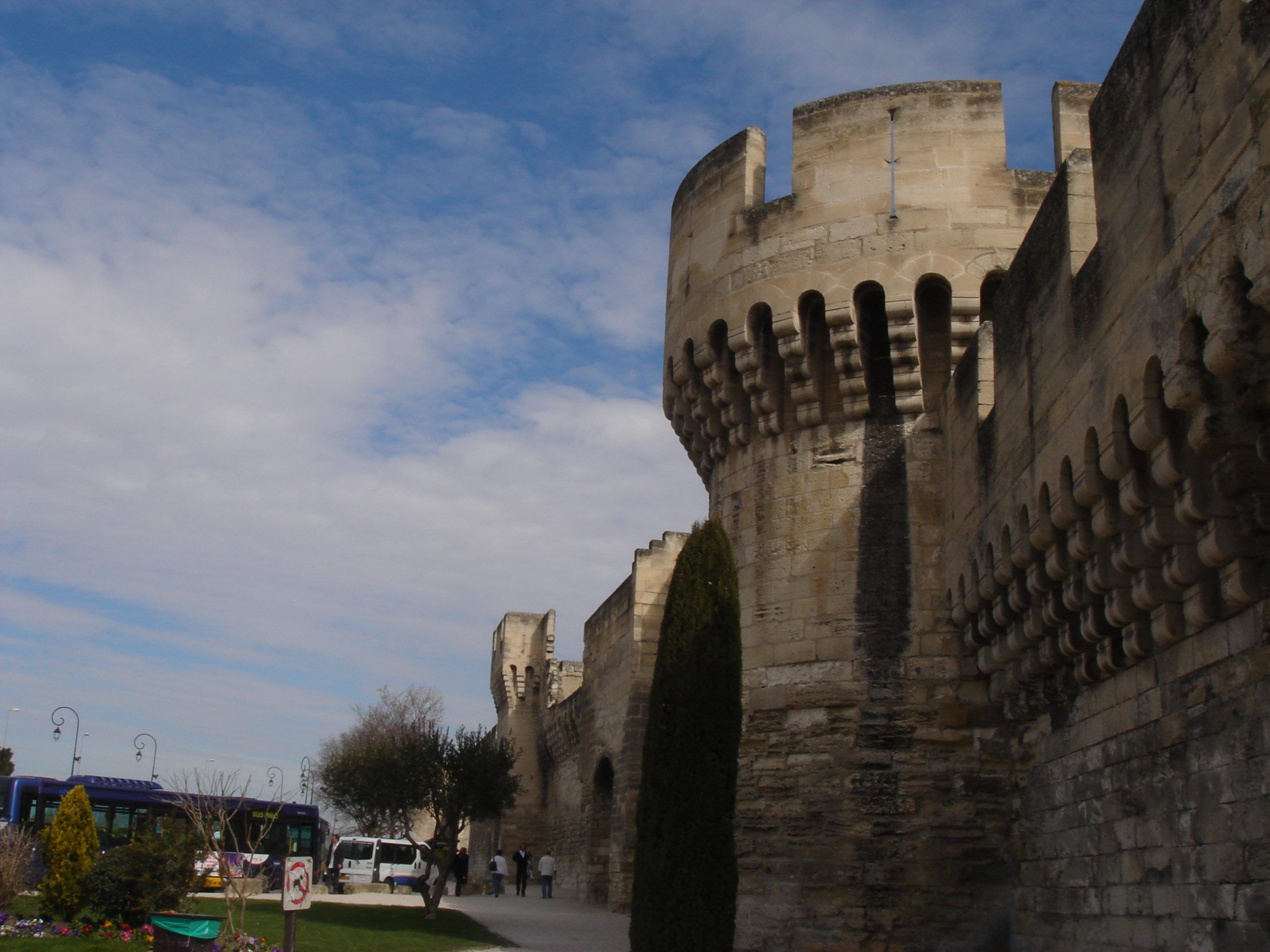 A section of the fortified walls of Avignon with crenellated towers and pedestrians walking nearby.