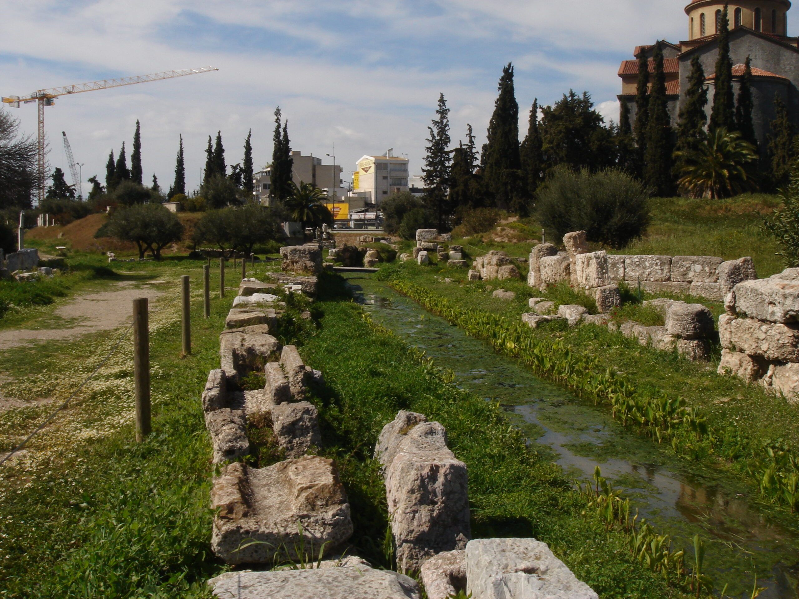 The Eridanos stream running through the Kerameikos, flanked by ancient stone blocks and bordered by cypress and olive trees.