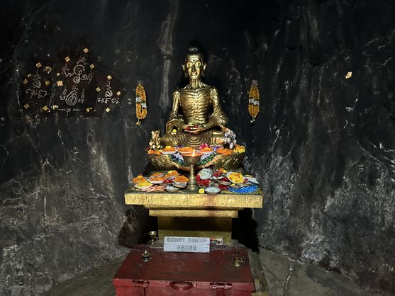 Gilded statue of the emaciated Buddha in a dark rock shrine at Dungeshwari Cave Temple, surrounded by offerings and garlands.