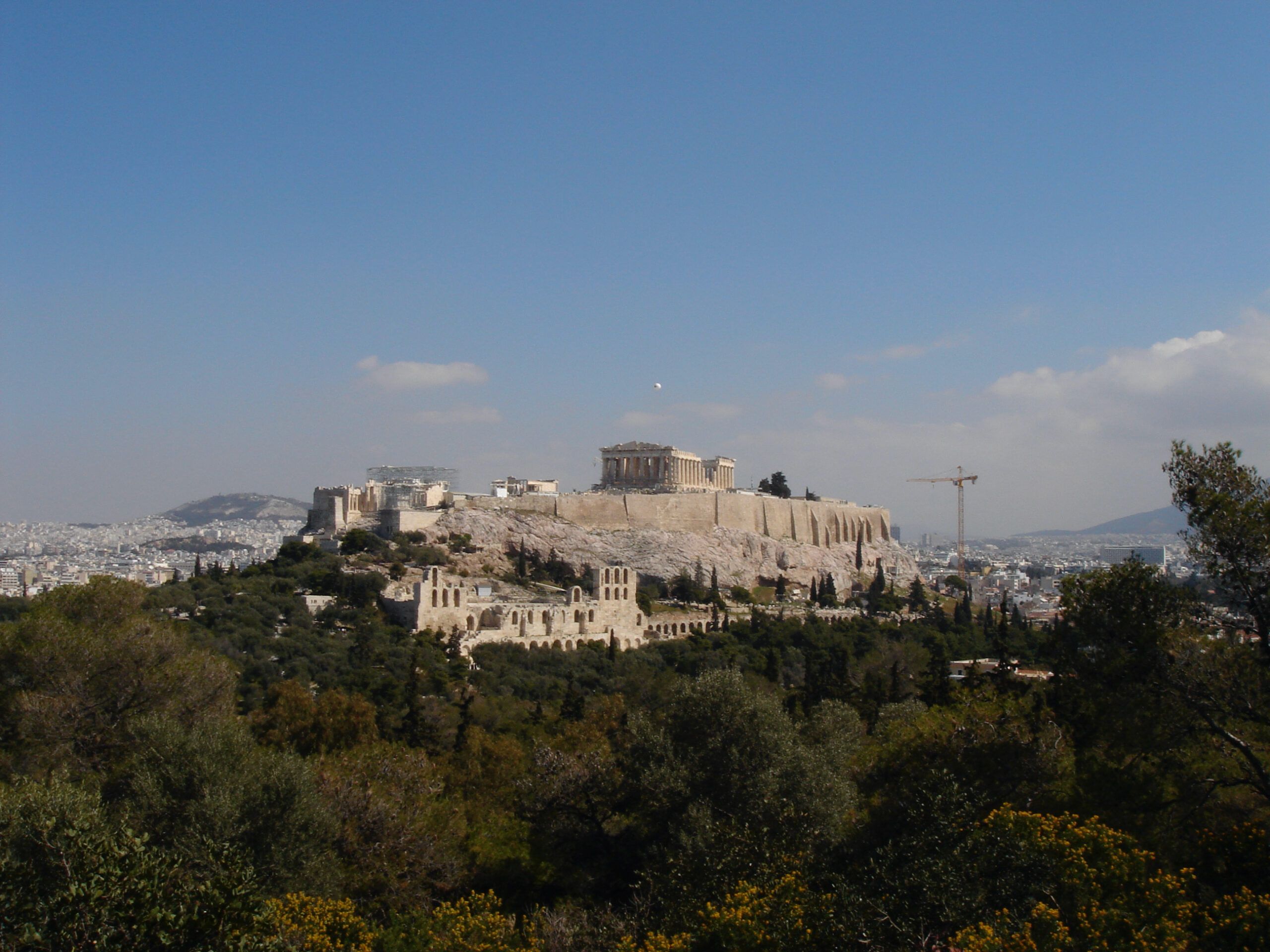 Panoramic view of the Acropolis in Athens, with the Parthenon and other ancient structures atop the rocky hill, surrounded by cityscape.