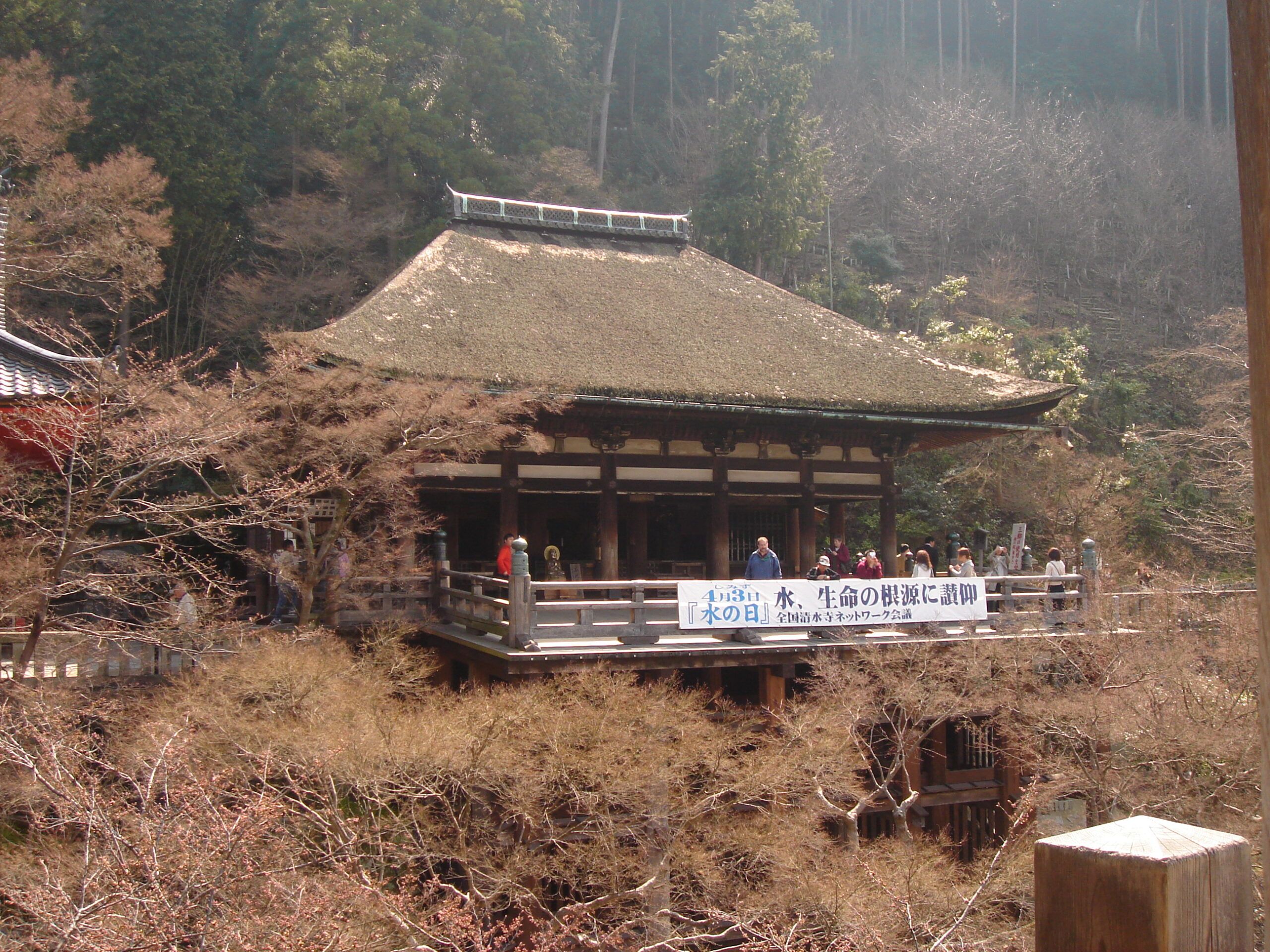 A hillside temple building with a thatched roof and a banner on the veranda, surrounded by forest and bare spring trees.