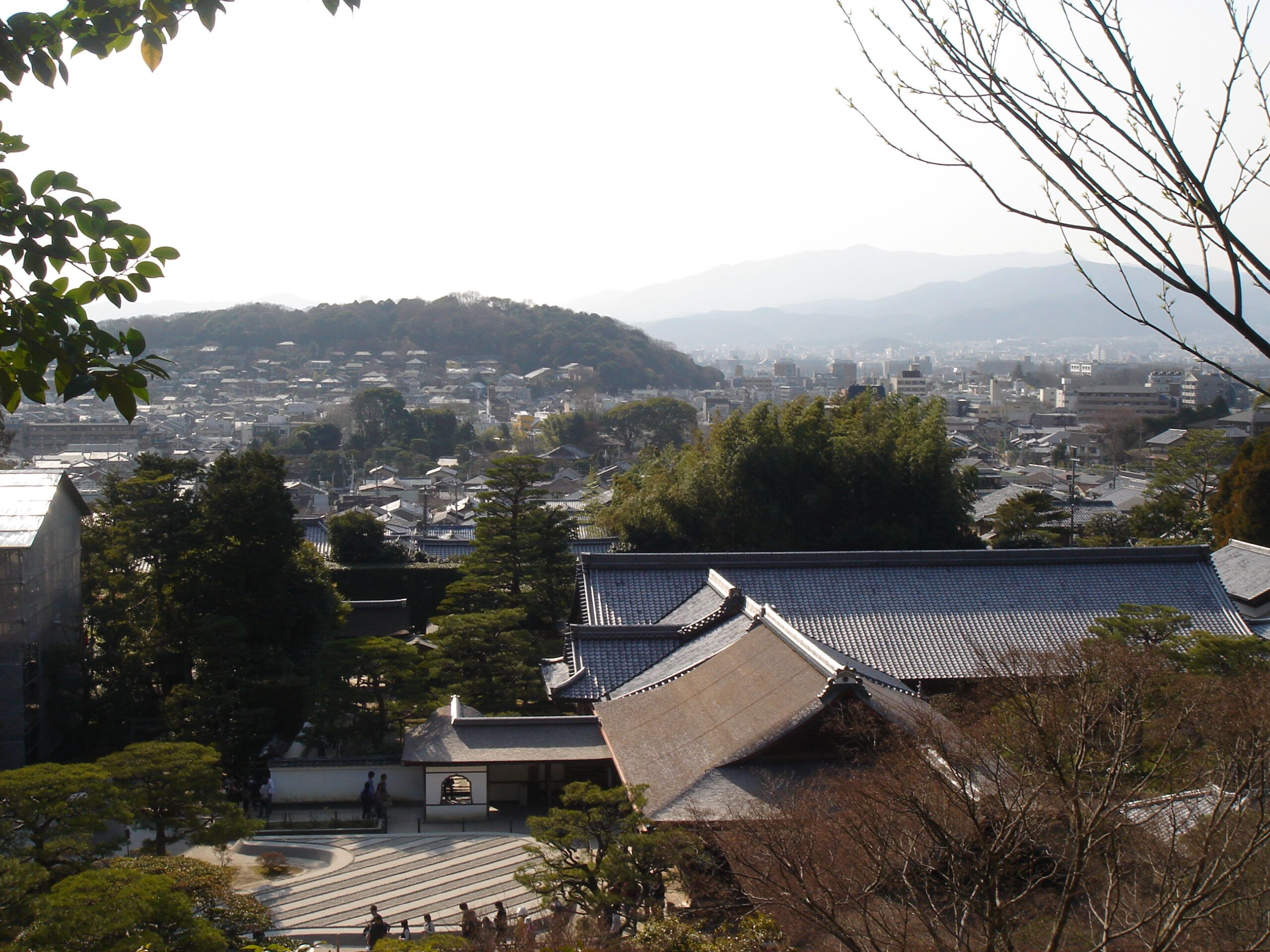 A panoramic view of Kyoto city with traditional temple rooftops in the foreground and mountains in the distance under a hazy sky.