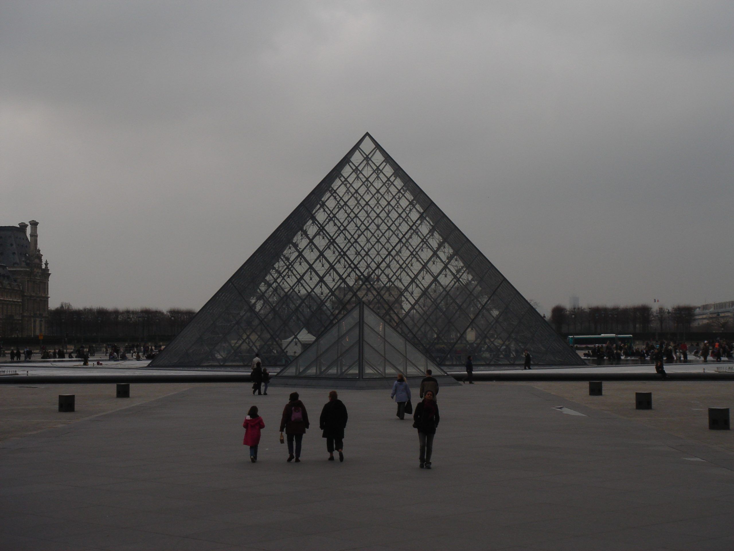 Wide view of the Louvre Pyramid in the Cour Napoléon with visitors walking in the courtyard.