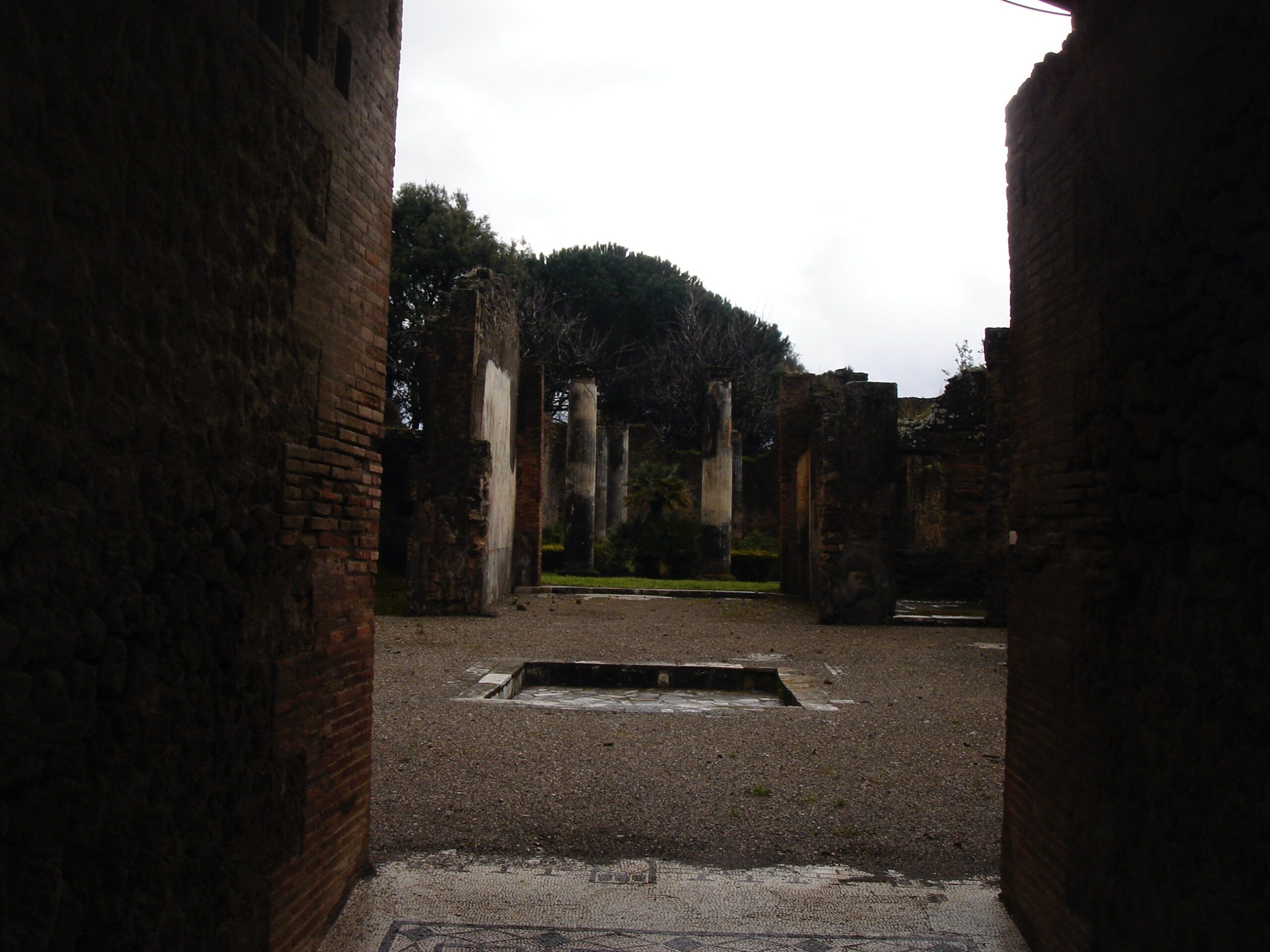 Shadowed view into a Roman atrium in Pompeii, looking toward a column-lined garden courtyard with a central impluvium.