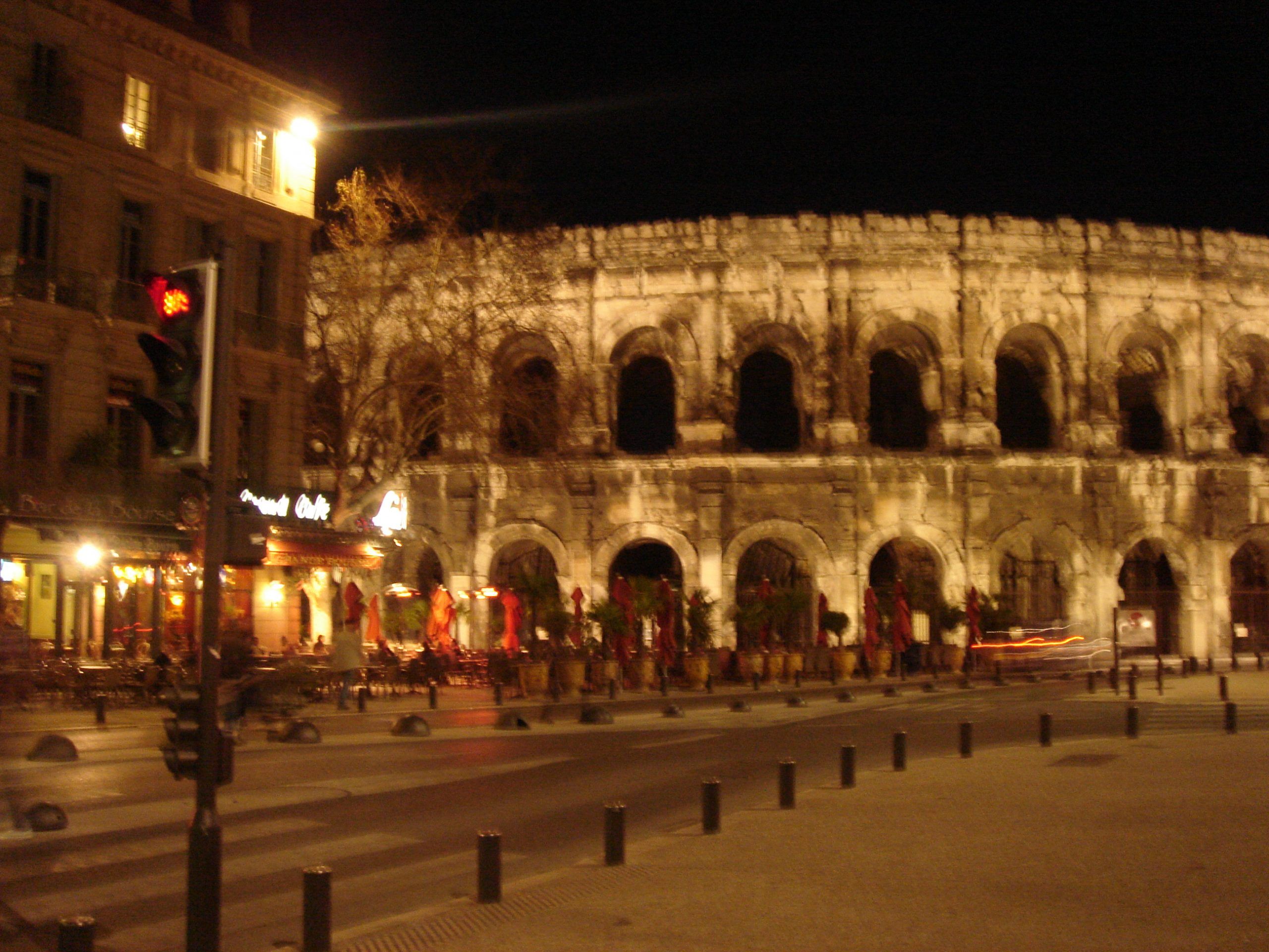 The Roman amphitheatre in Nîmes illuminated at night, with a restaurant and traffic lights in the foreground.