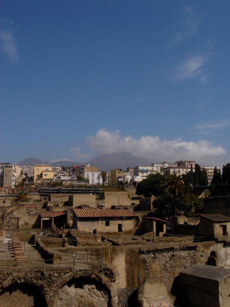 View over the ruins of Herculaneum with modern buildings and Mount Vesuvius visible in the background under a blue sky.