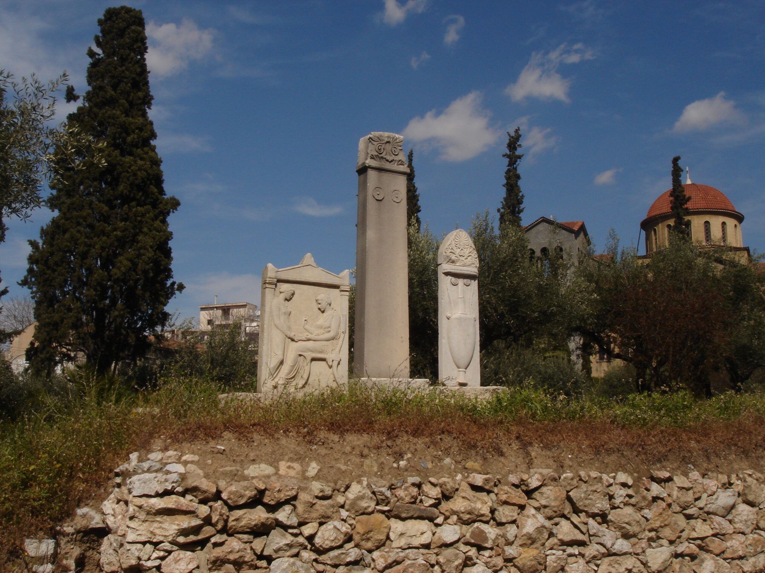 Three marble funerary stelae from classical Athens, including a sculpted relief of two seated figures, set in a peaceful grove with cypress trees and a red-domed church.