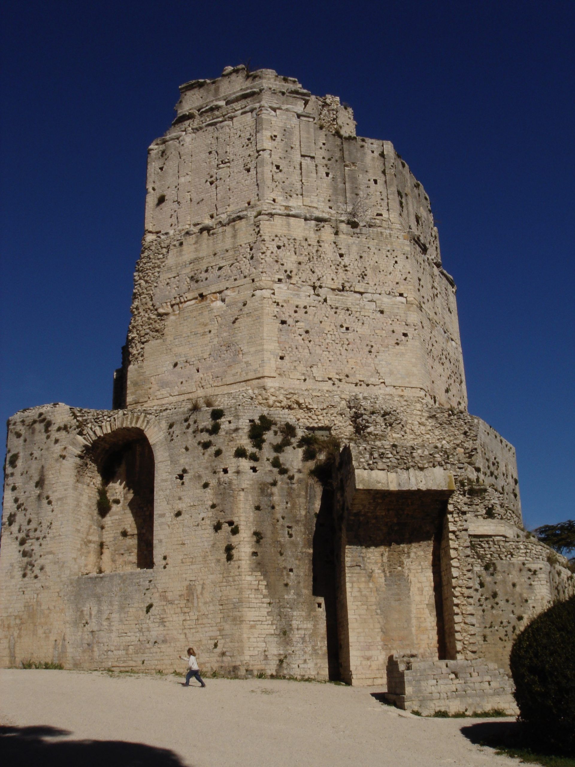 A Roman-era stone tower under bright sun, with a small child walking nearby for scale.