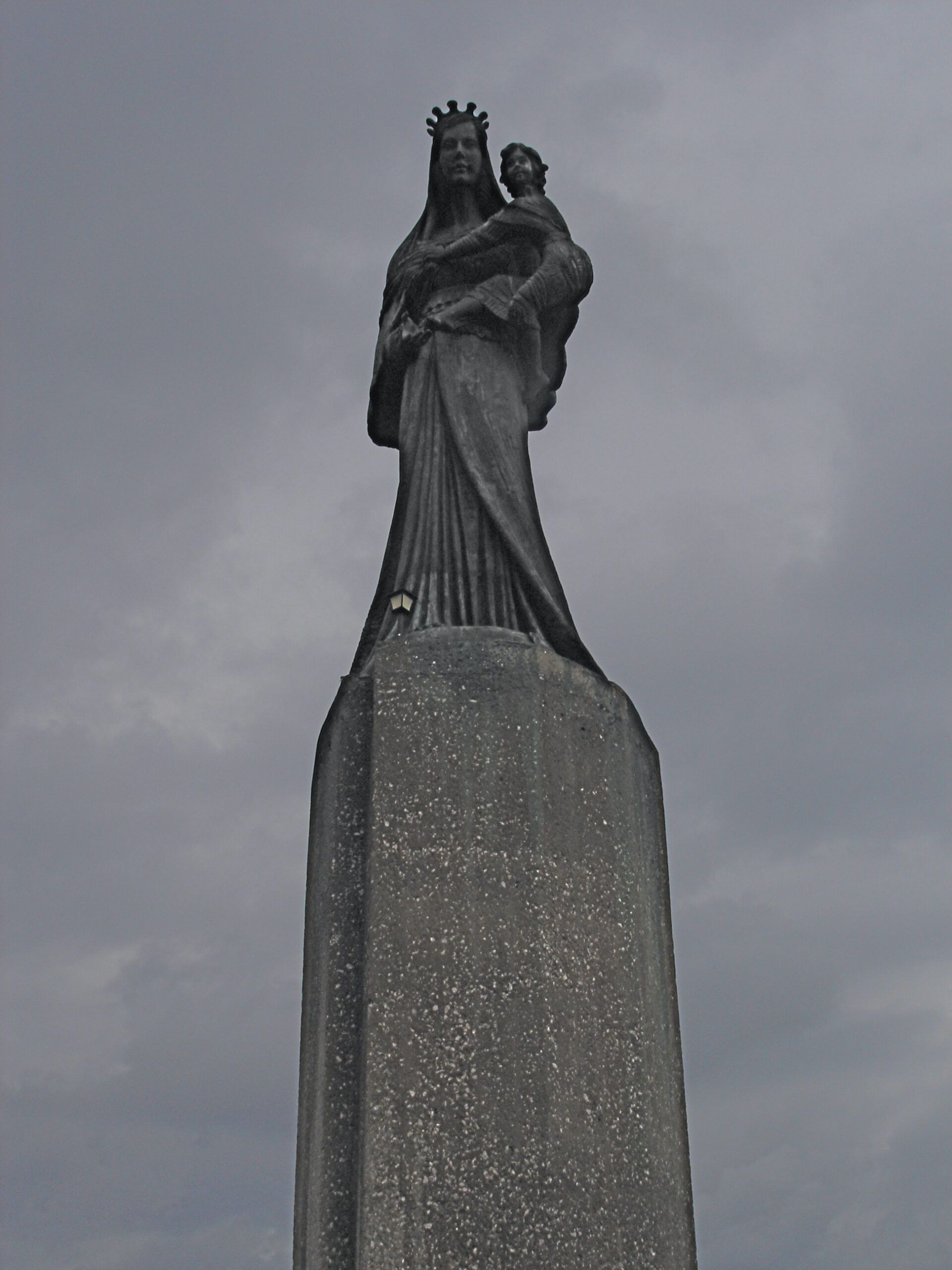A dark bronze statue of the Virgin Mary holding the infant Jesus, mounted on a tall pedestal and silhouetted against a cloudy sky.