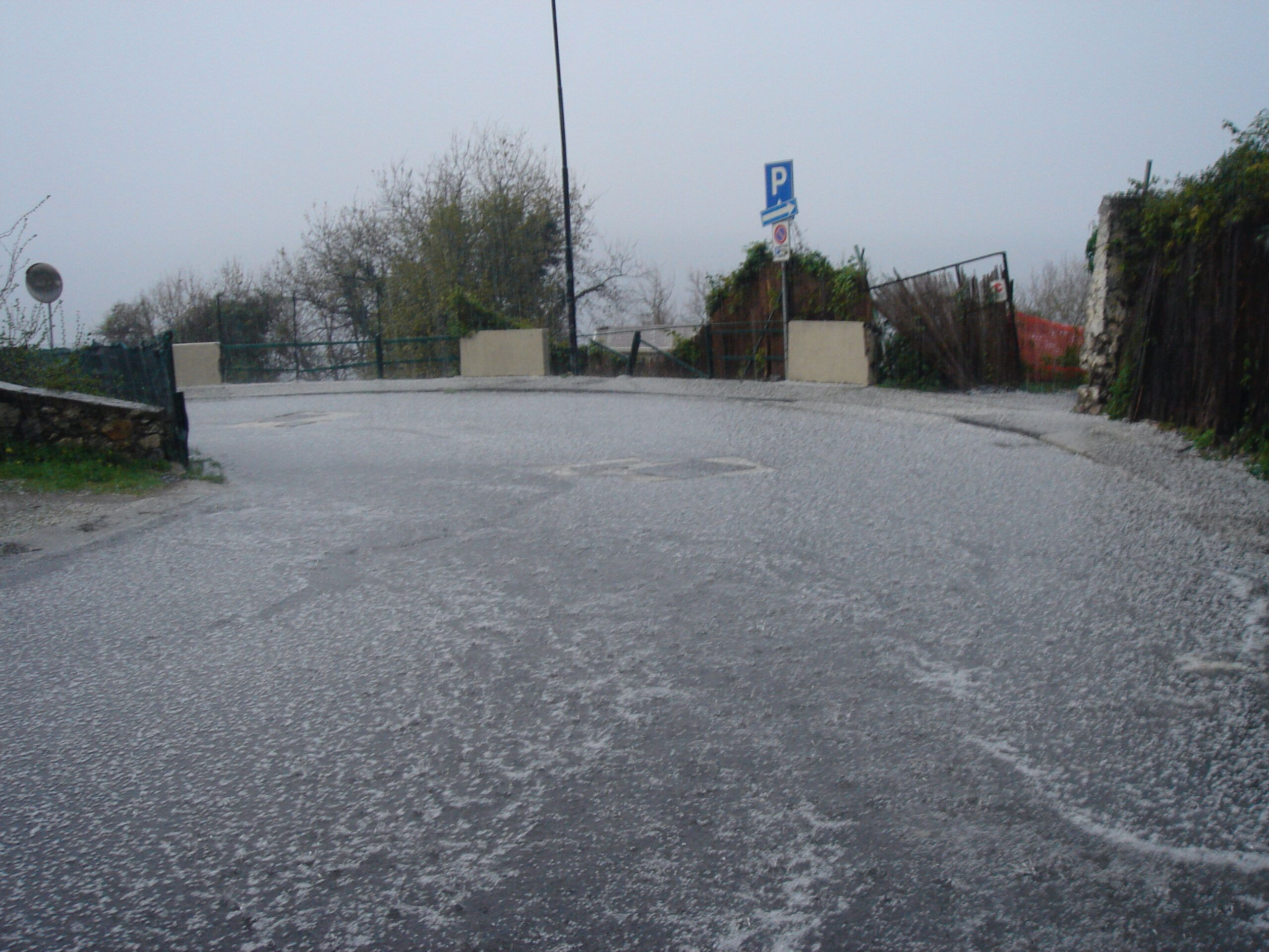 A steep road blanketed in hail, with broken gates and a parking sign in the background, under heavy grey skies.