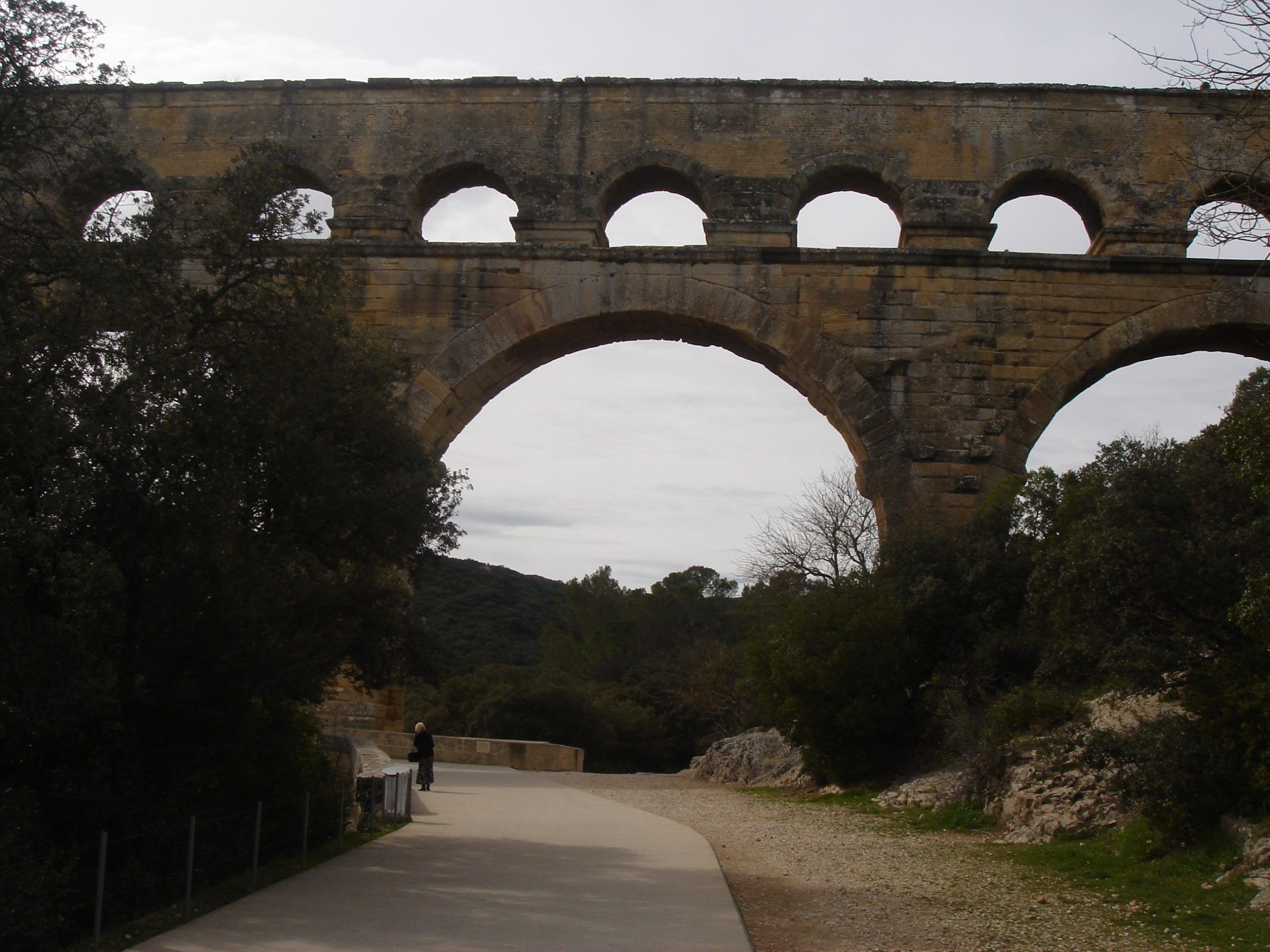 View of the Pont du Gard from below, showing a large central arch and upper tiers of the aqueduct framed by trees and a walking path.