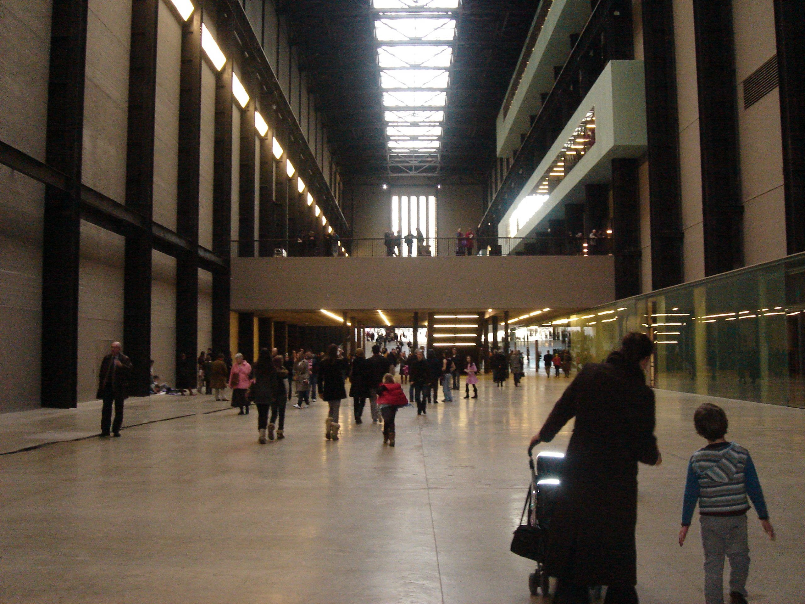 A massive gallery, stories tall, with some people milling around in the distance. Angela and the kids continue to head away from us.
