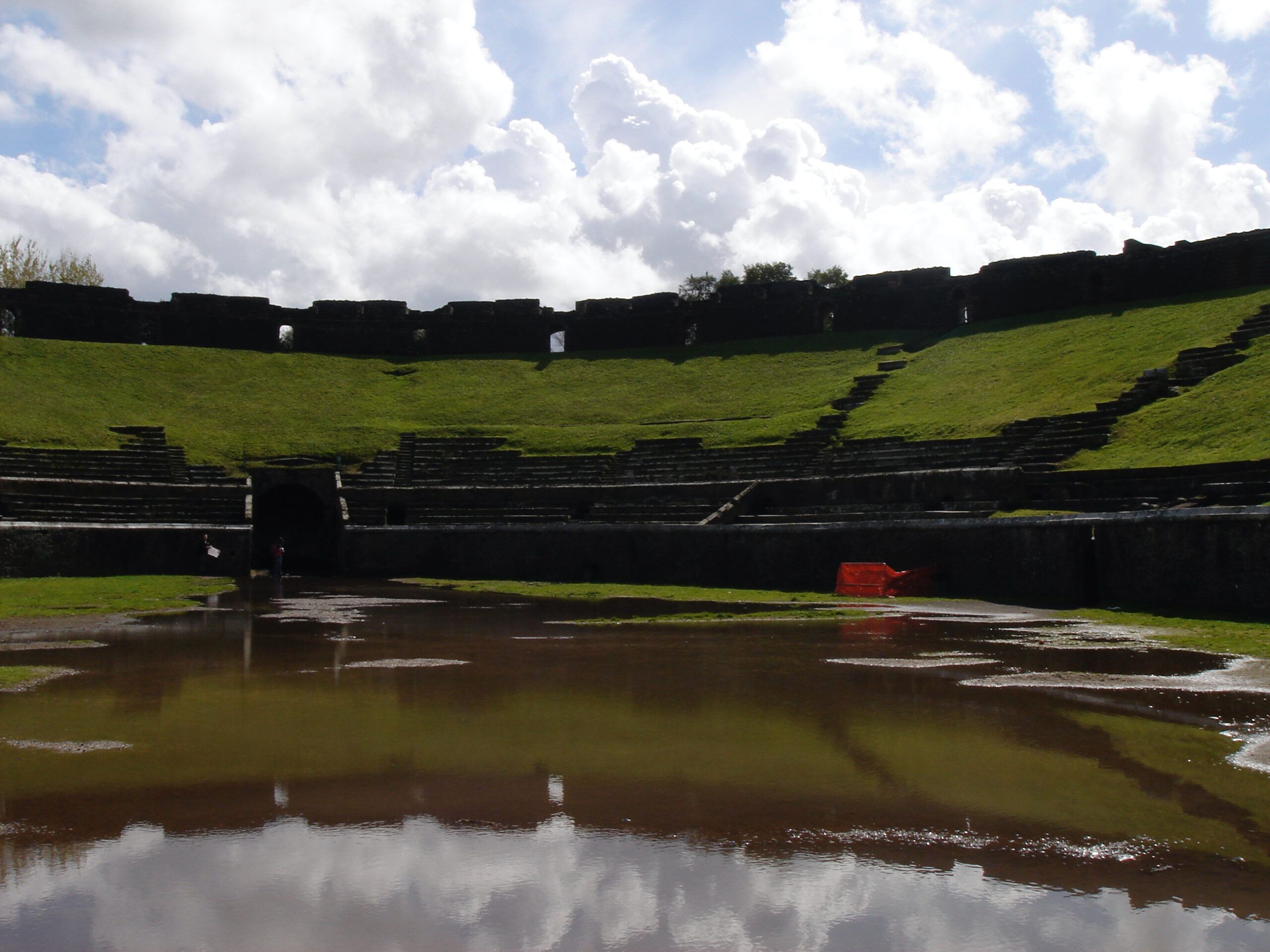 The amphitheatre at Pompeii, its arena flooded with rainwater and framed by steep green embankments.