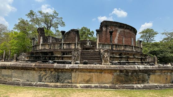 The ruins of the Polonnaruwa Vatadage, a circular brick shrine with ornate stone steps and a seated Buddha statue at its center, framed by crumbling columns and lush greenery.