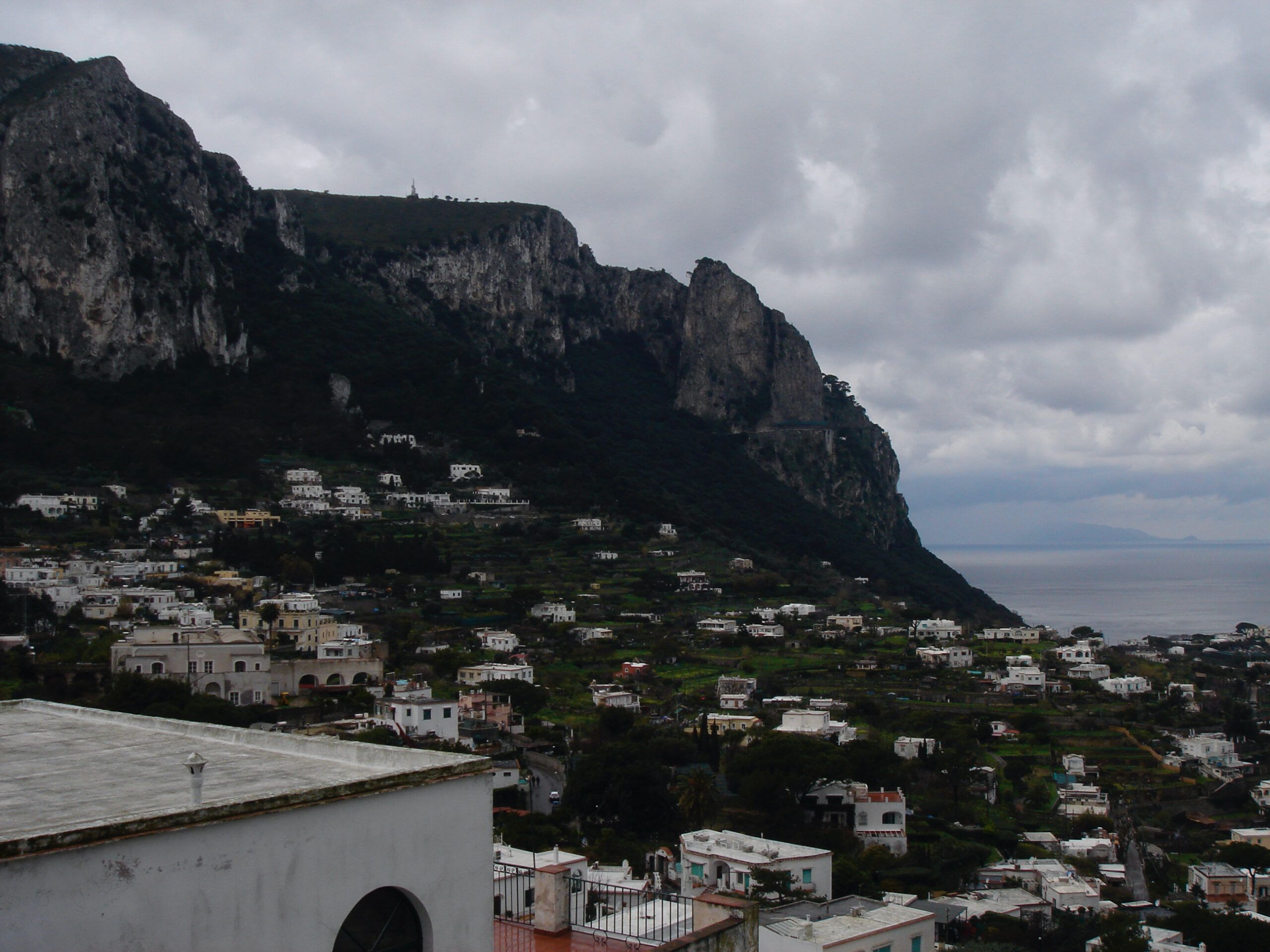 View of a coastal town with white buildings spread across a green hillside, under a dramatic cloudy sky and steep cliffs.