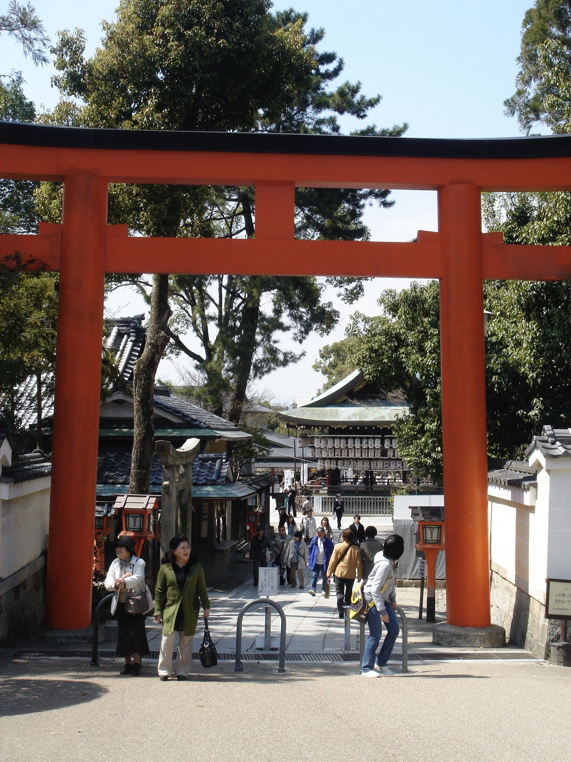 A large red torii gate at the entrance to a temple complex, with visitors walking along a paved path lined by lanterns and walls.