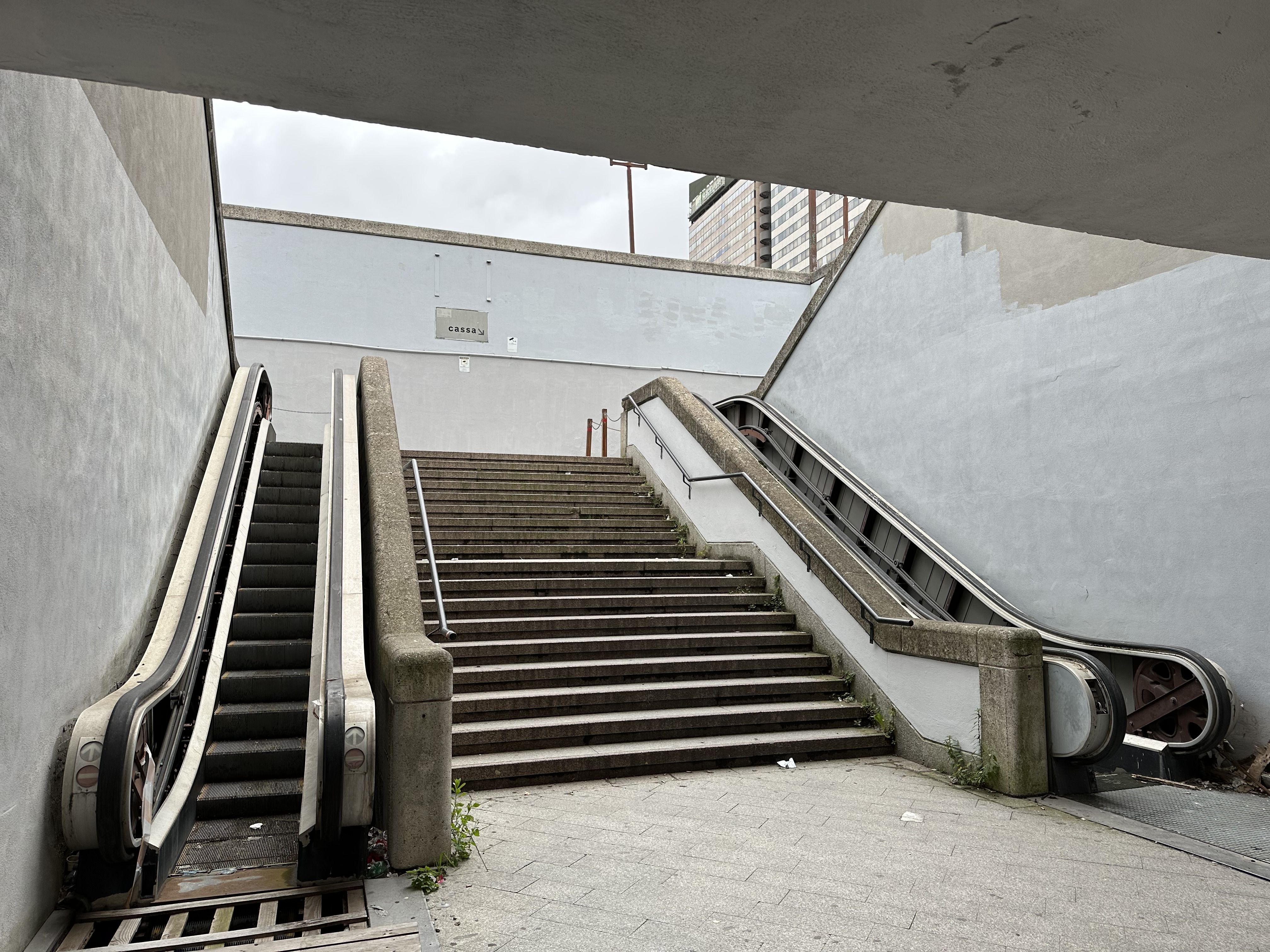 View from below of a stairway flanked by two broken escalators in a concrete underpass, with weeds growing between the steps.