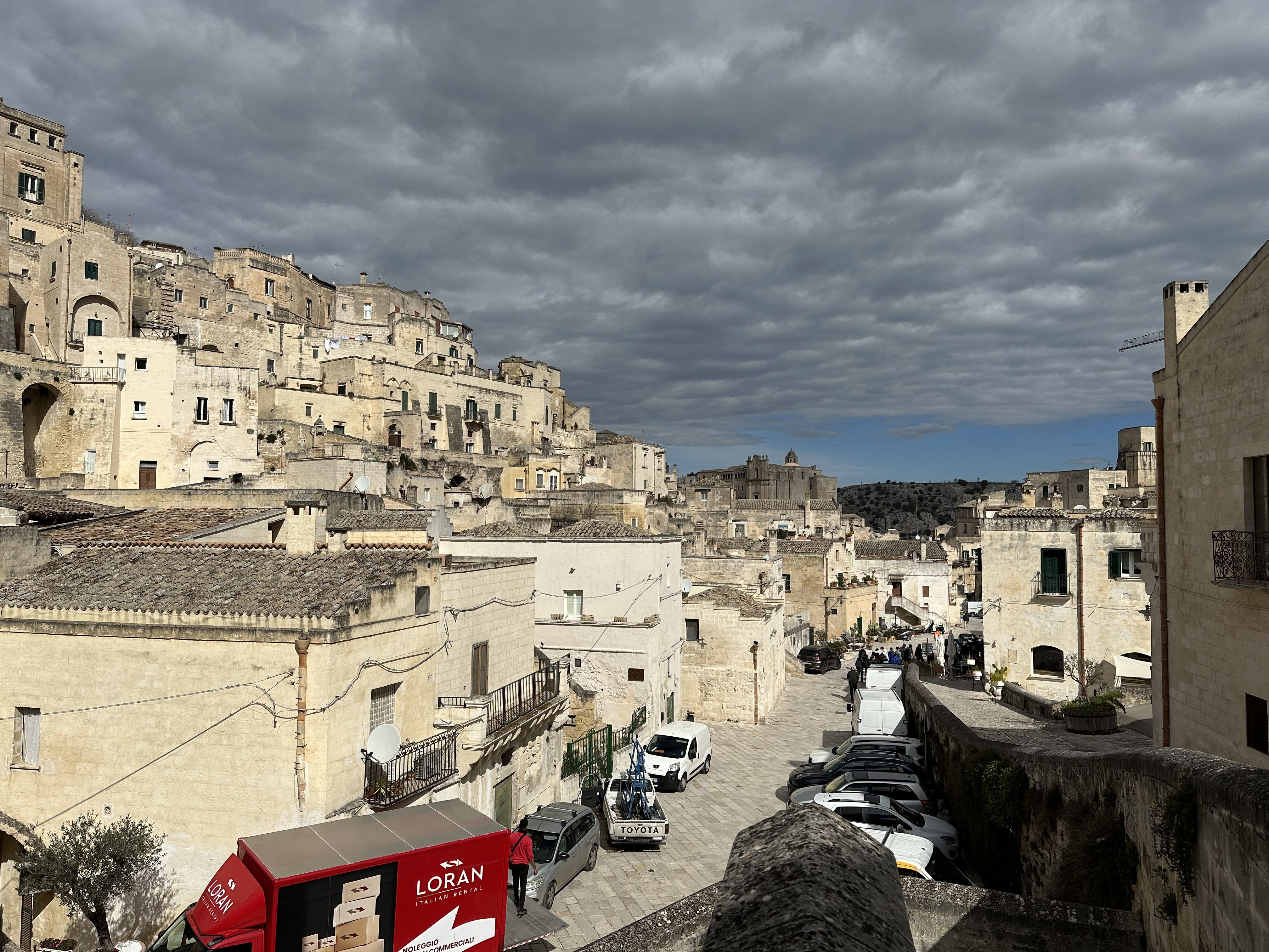 Cluster of stone buildings in Matera’s Sassi district, with cloudy skies and a red delivery van in the foreground.