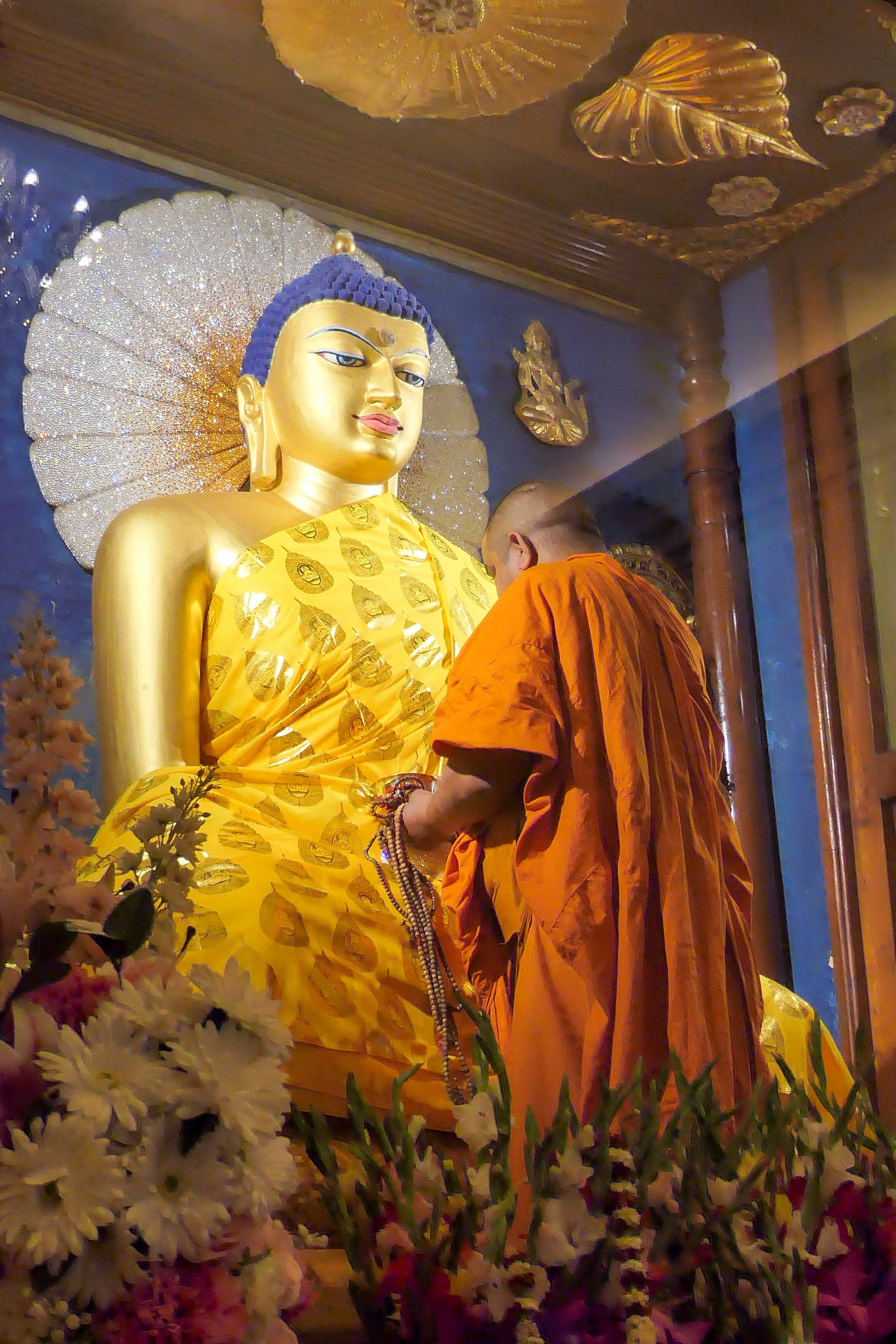A monk in orange robes adorns a large golden statue of the Buddha with prayer beads inside a shrine.