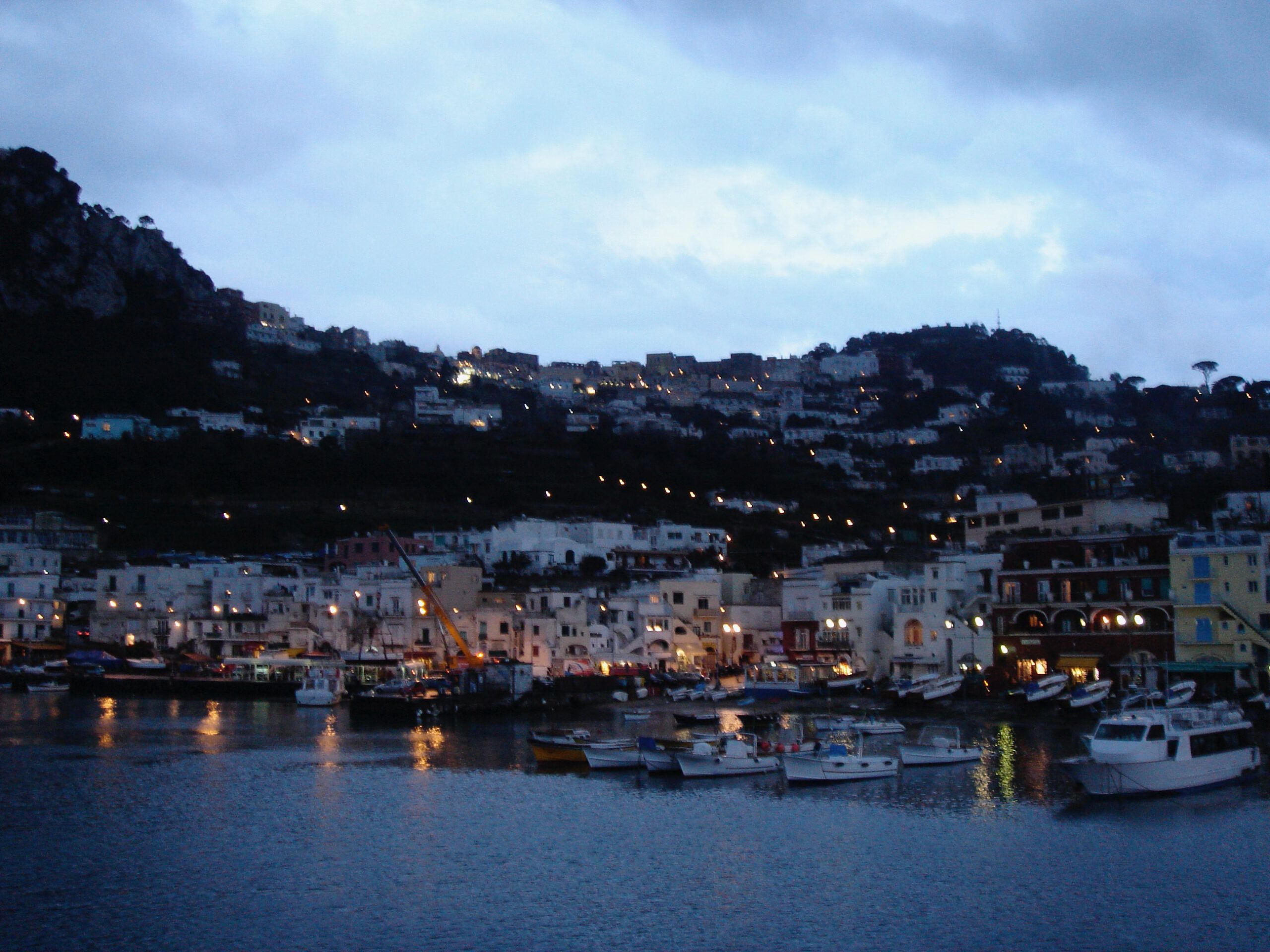 The harbour at Porto Grande on Capri at nightfall, with fishing boats in the foreground and the hillside town lit up in the background.