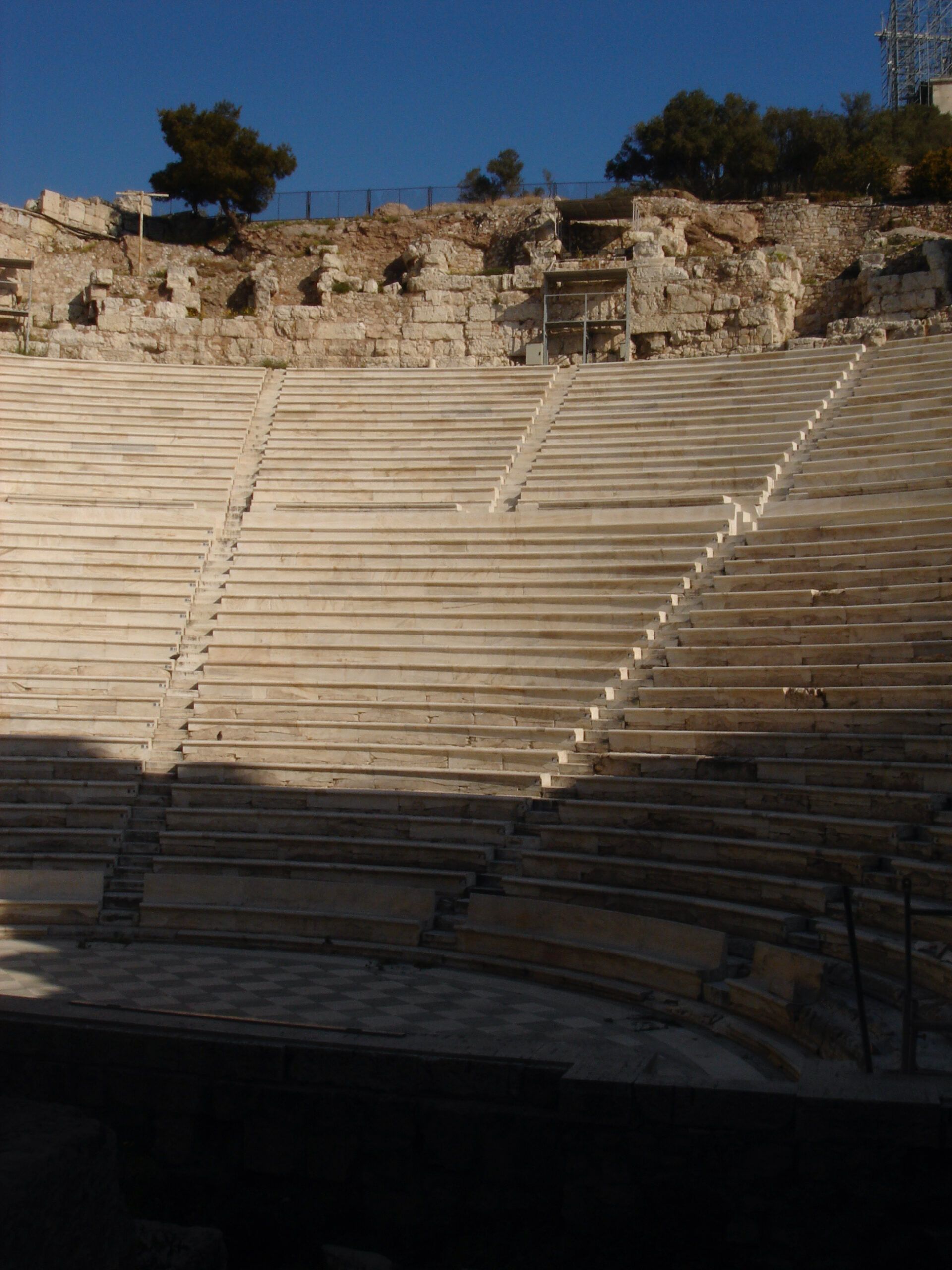 Wide view of an ancient stone amphitheatre with concentric seating tiers and partial ruins on the hillside.