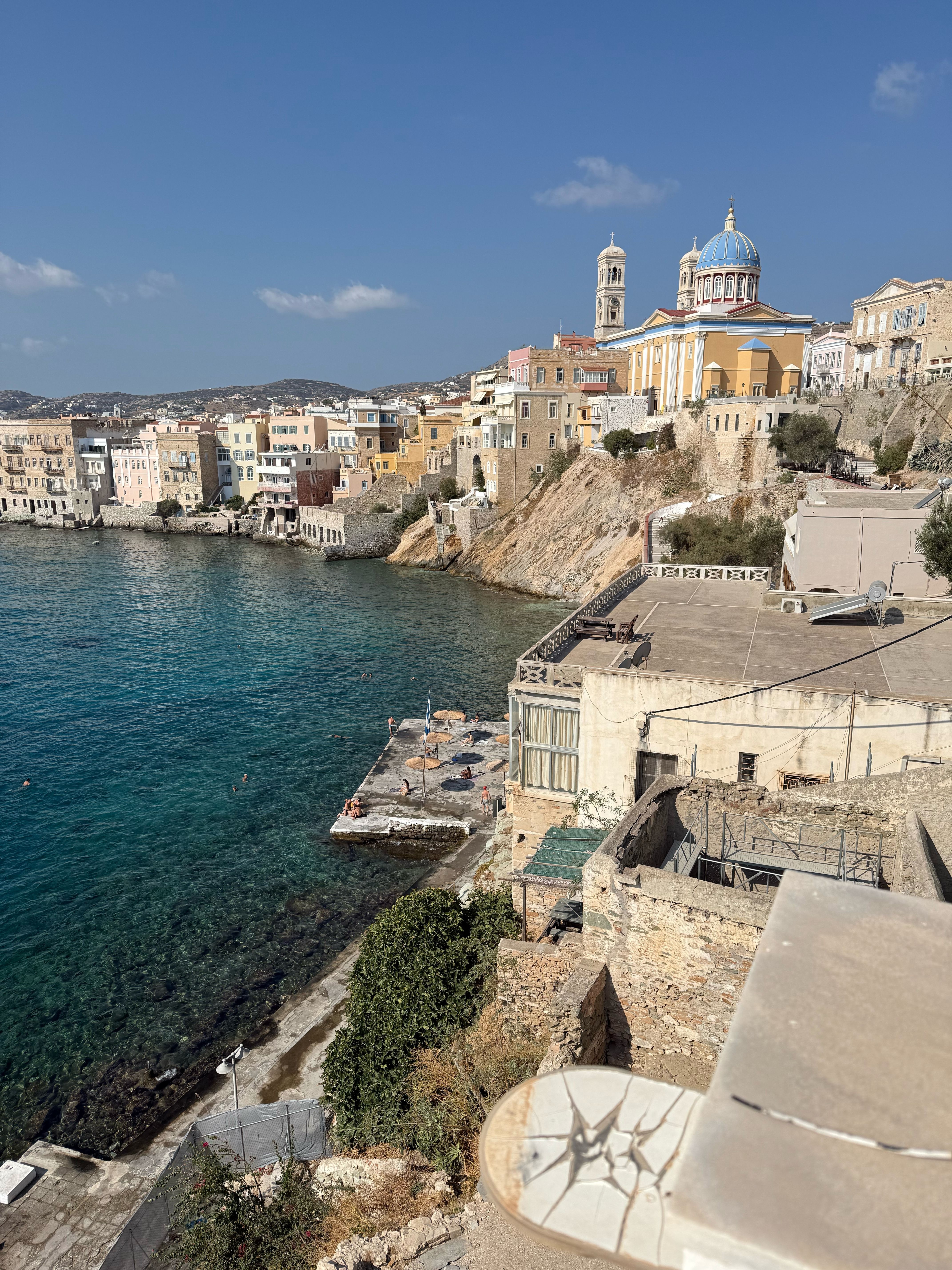 Looking down from the hotel terrace. The water is deep blue, and a concrete jetty with beach umbrellas juts out into it. Above the jetty are buttressed stone walls, and far above them is the domed church of Saint Nicholas.