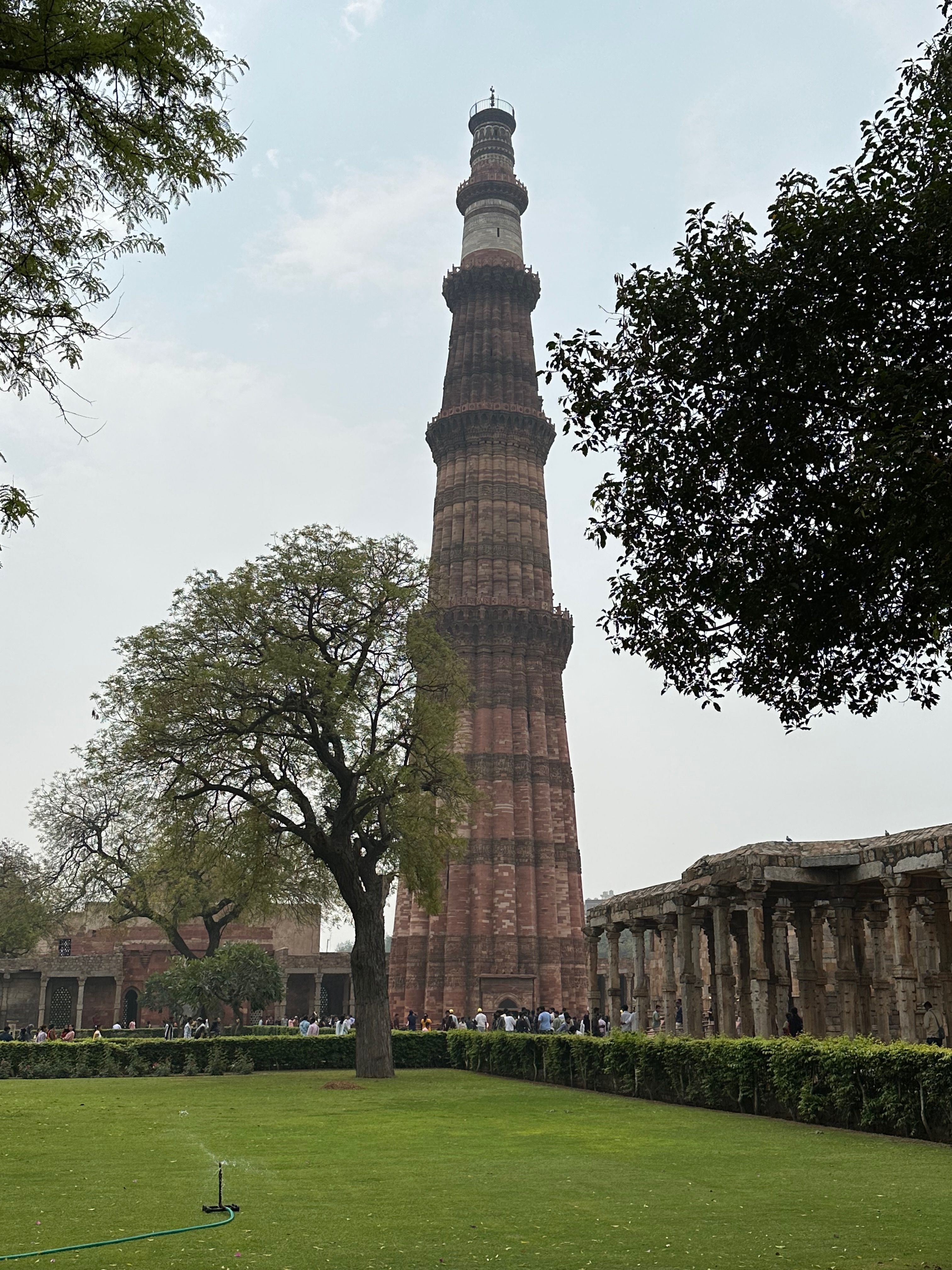 The Qutb Minar rising above the trees and surrounding ruins, seen from a well-kept lawn with visitors at its base.