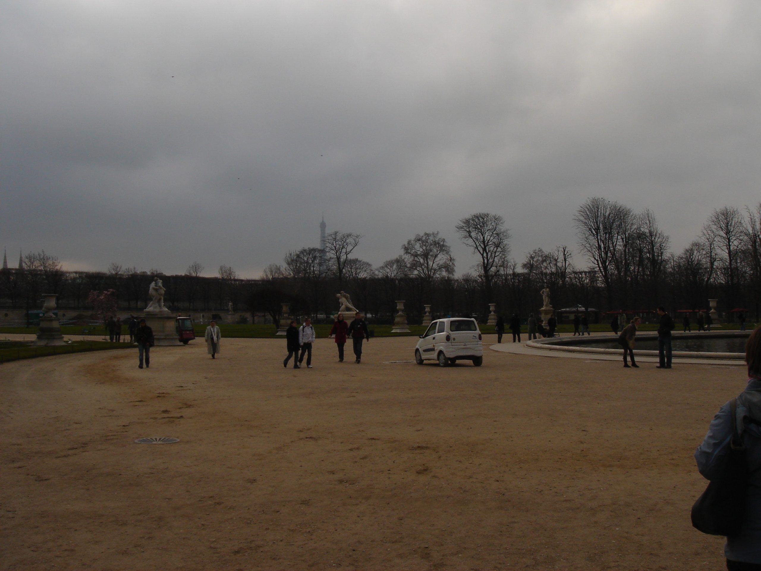 People walking in the Jardin des Tuileries with statues, a pond, and the Eiffel Tower in the distance.