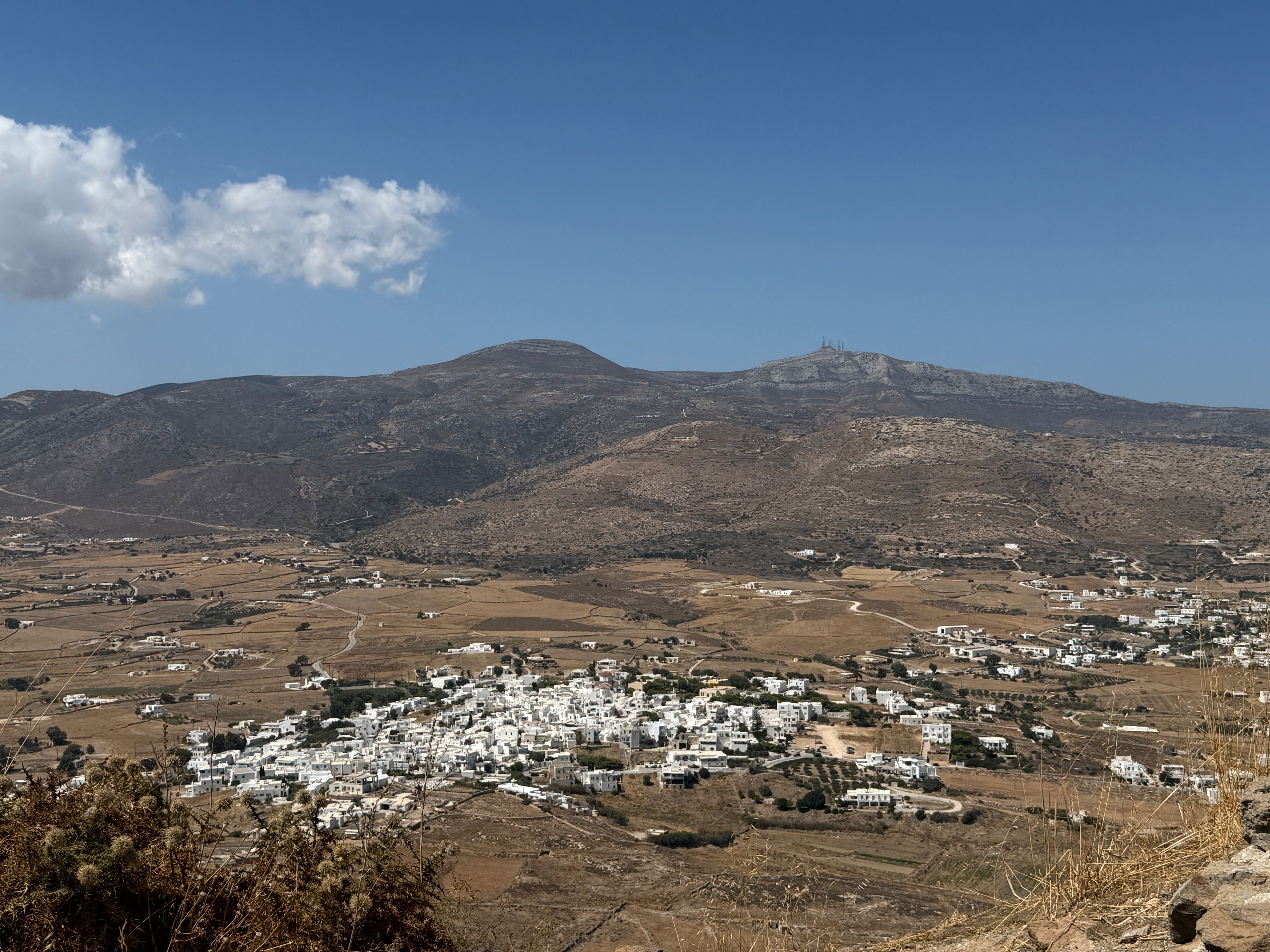 A view from the hilltop monastery of Agios Antonios, looking out over a dry valley. There is a village made up of white buildings and a mountain in the distance; the sky is blue with one or two small clouds.