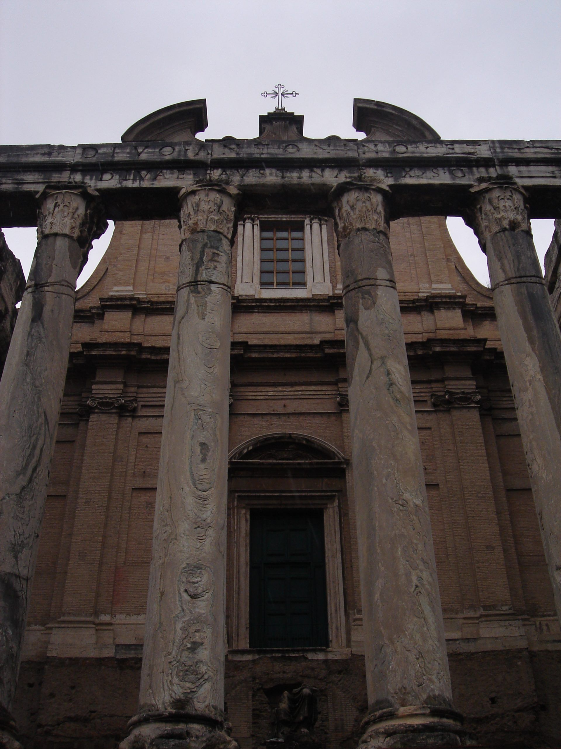Low angle shot of the Temple of Antoninus and Faustina, highlighting its tall Corinthian columns and baroque church facade behind them.