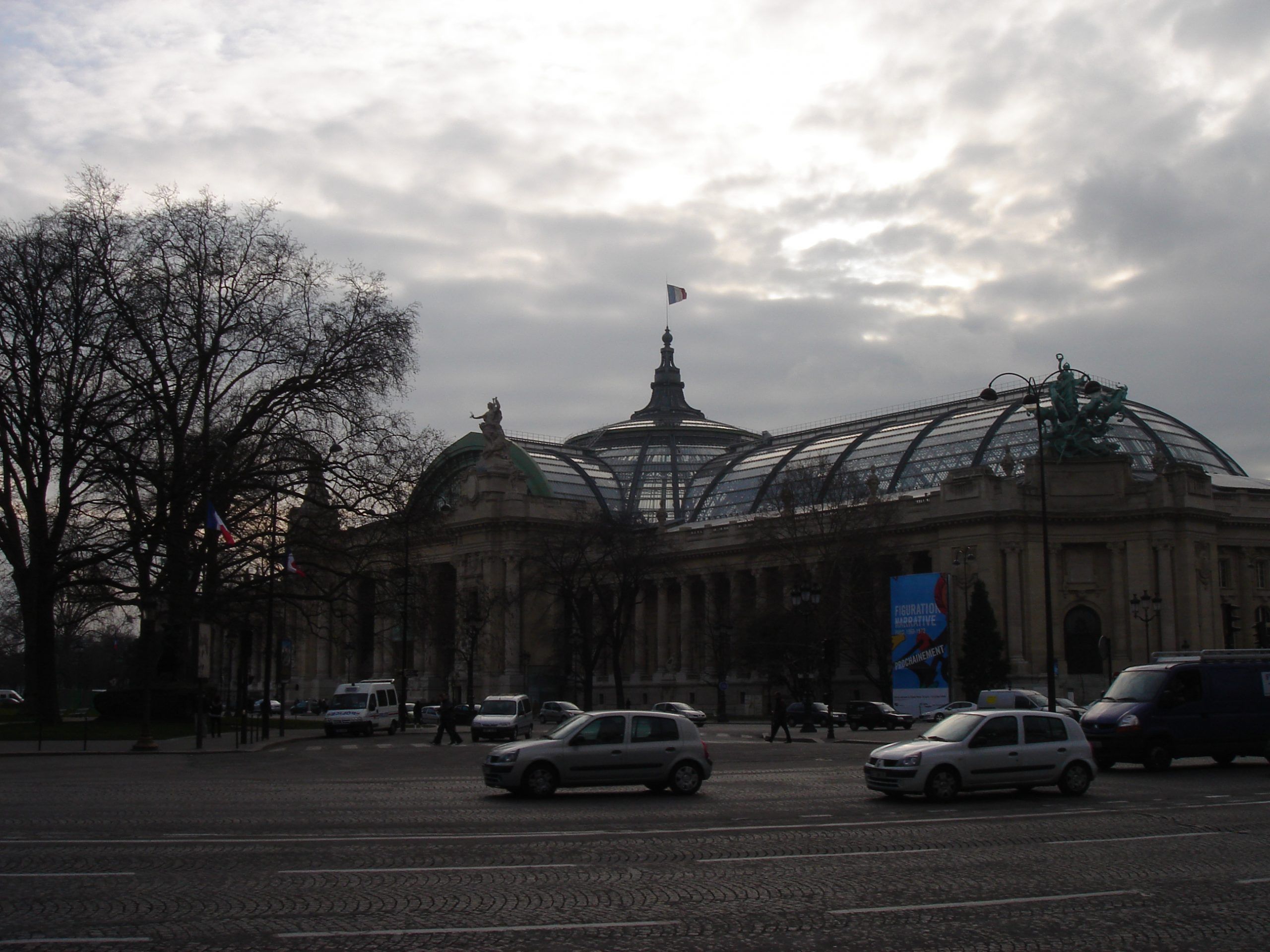 Facade of the Grand Palais in Paris with glass dome and French flags, seen from across a busy street.