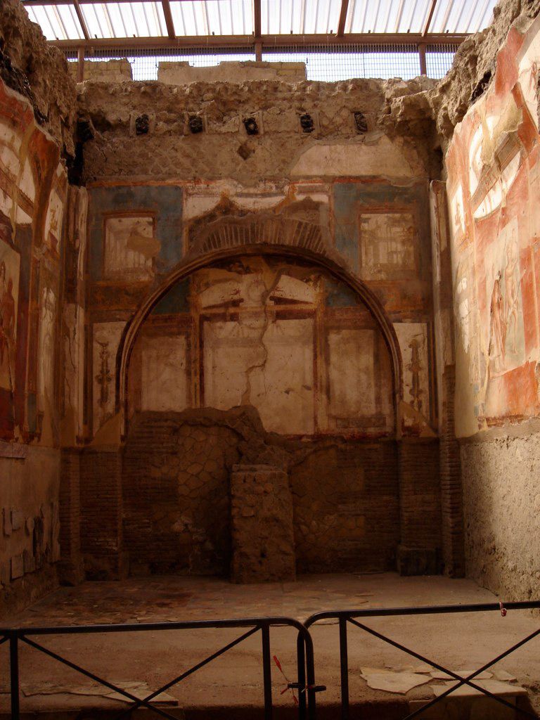 Frescoed room in Herculaneum with vivid red and blue wall paintings, partially damaged but showing architectural motifs and niches.