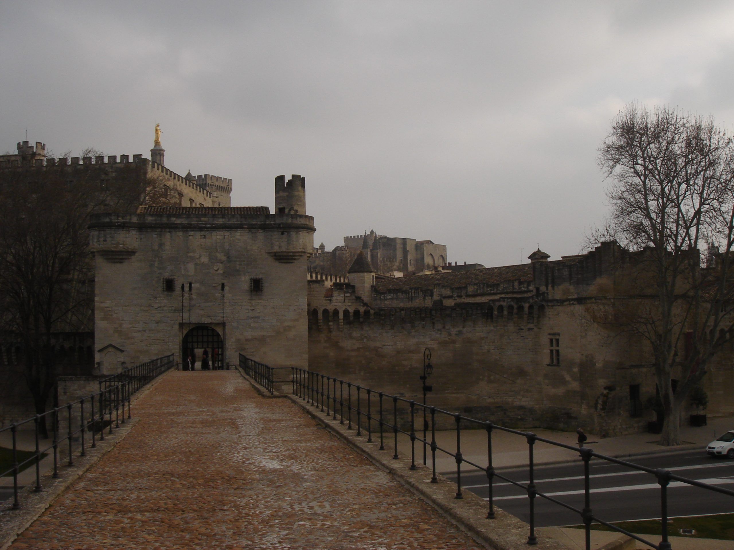 View across a bridge leading to the entrance of the Papal Palace in Avignon, with the golden statue of the Virgin Mary visible in the background.