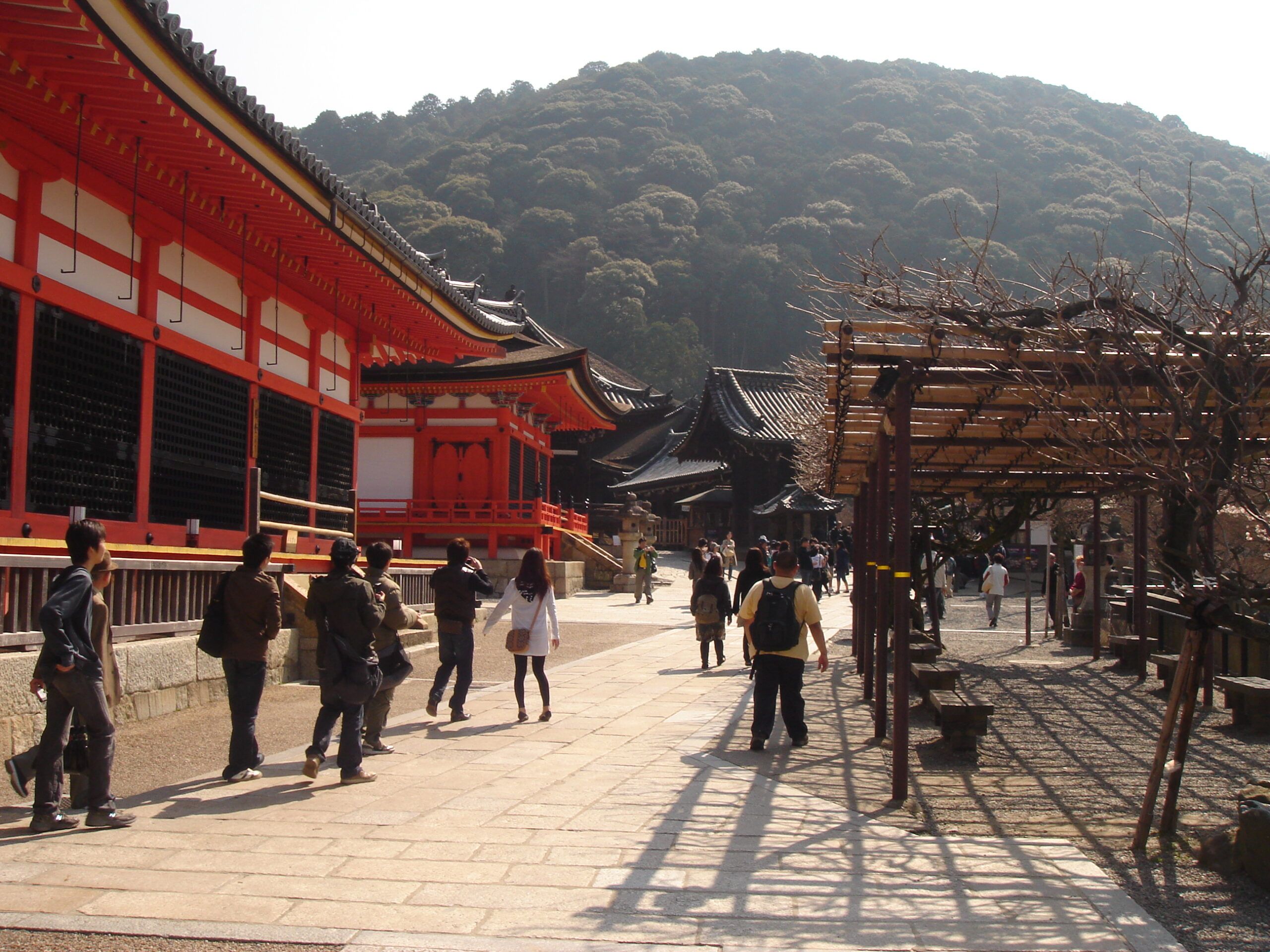 Visitors walk through a wide open temple complex lined with vivid red-and-white buildings and trellises under a forested hill.