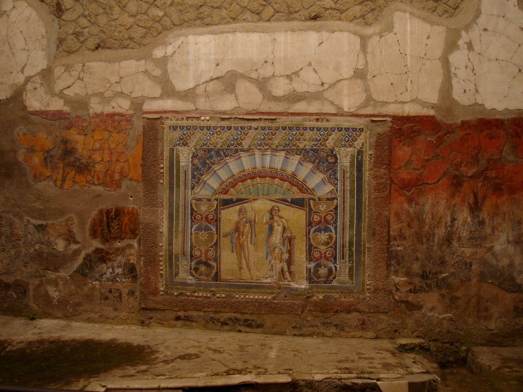 Brightly colored wall mosaic in Herculaneum depicting a small aedicula scene with two standing figures beneath a decorative arch.