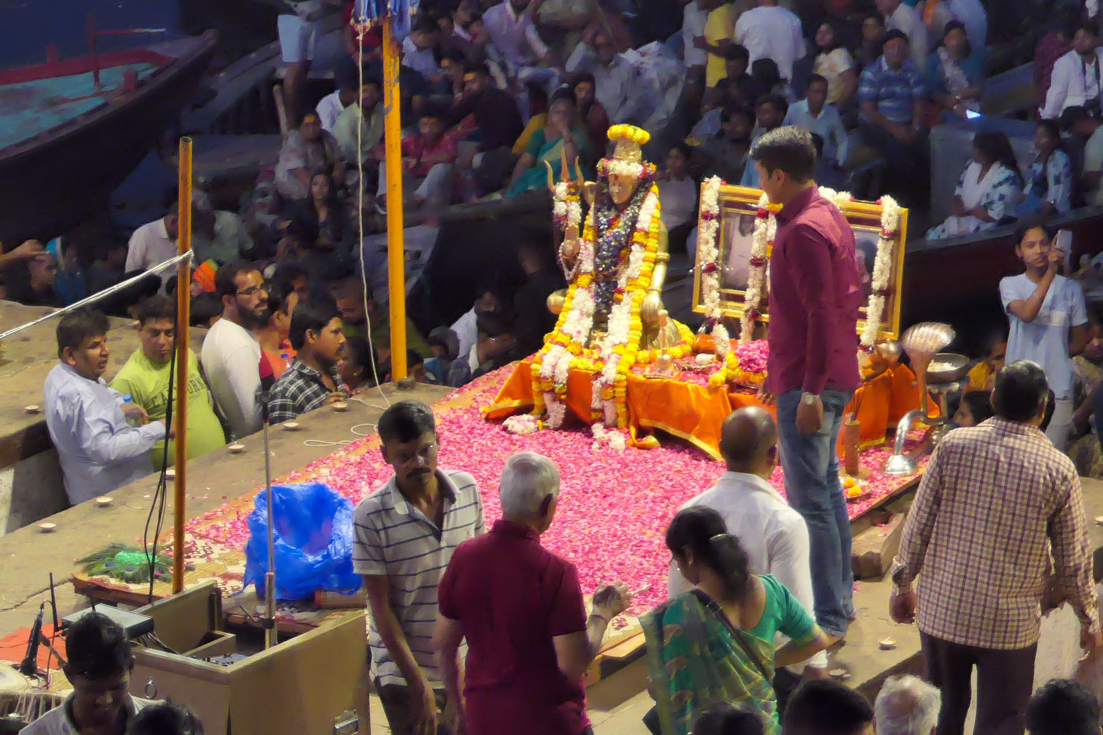 Close-up of a flower-covered platform with a statue of a deity at Dashashwamedh Ghat, surrounded by people preparing for the Ganga Aarti ceremony.