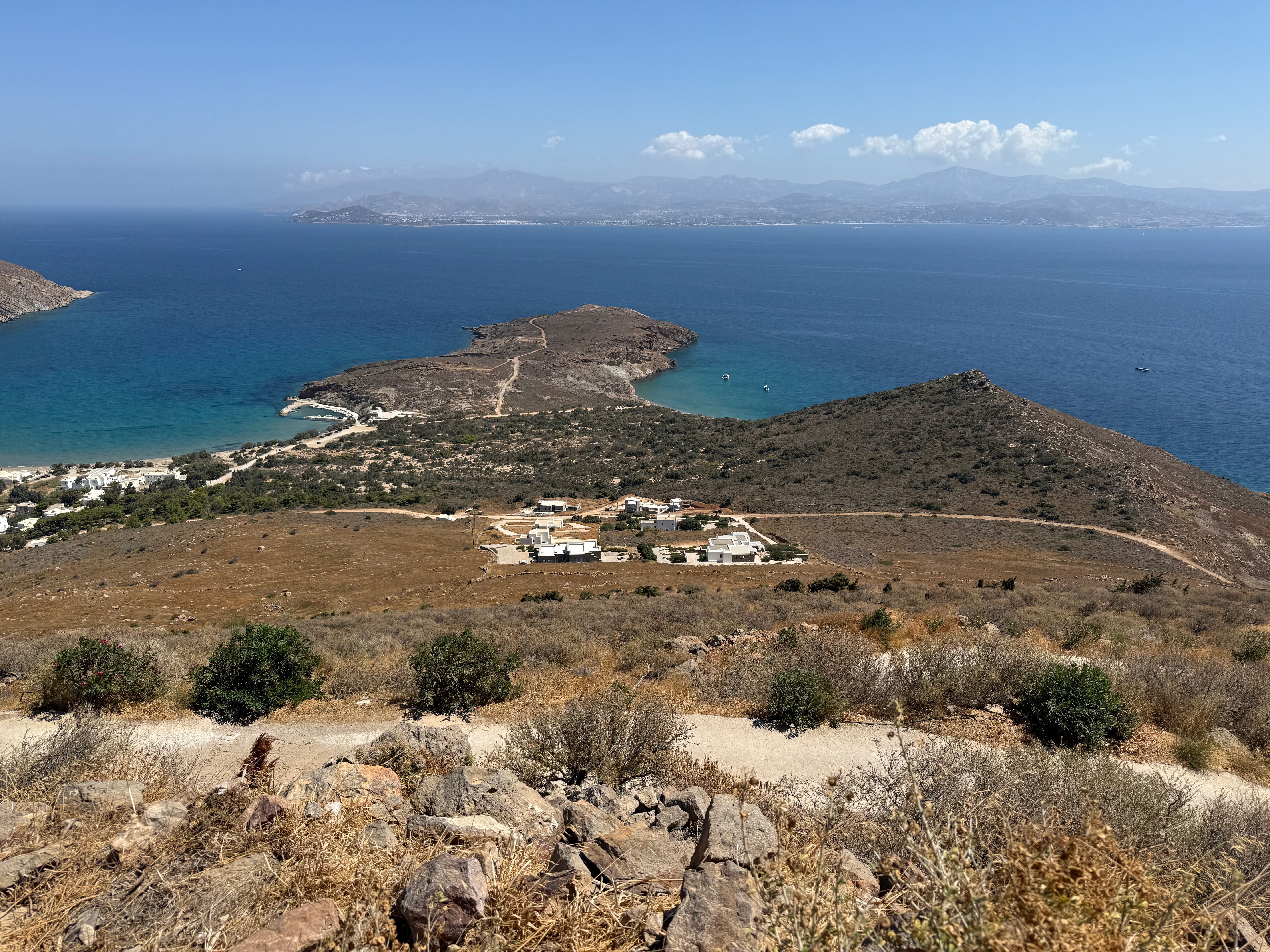 Another view from Agios Antonios. A dry promontory juts out into the sea, with a small white chapel on its tip. There are a few white buildings in the middle distance and some buildings near the shore on the left. The sea is deep blue. In the distance is the island of Naxos is hazily visible.