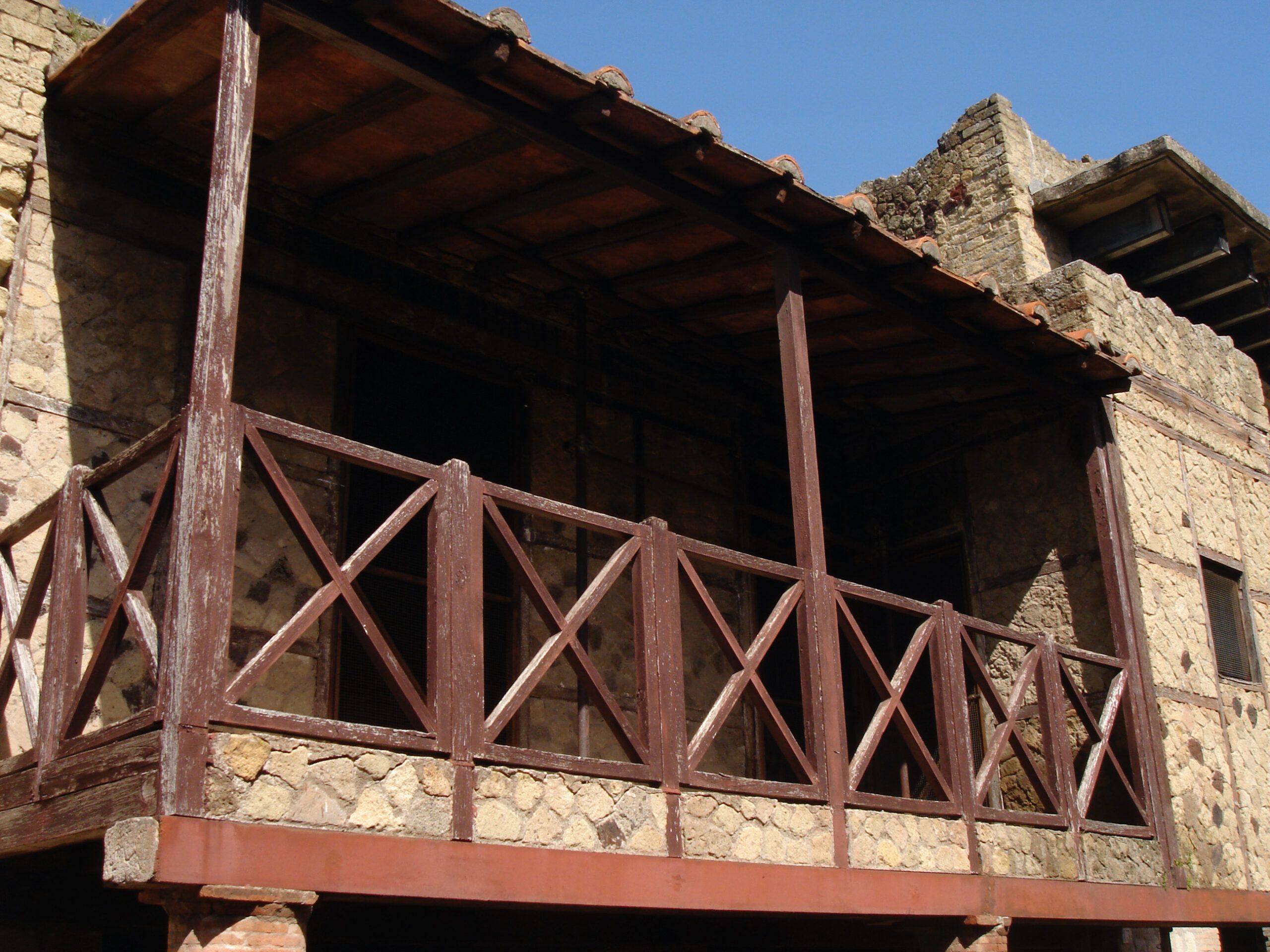 Wooden balcony on the upper level of a Roman house in Herculaneum, reconstructed with cross-braced railings and supported by brick columns.