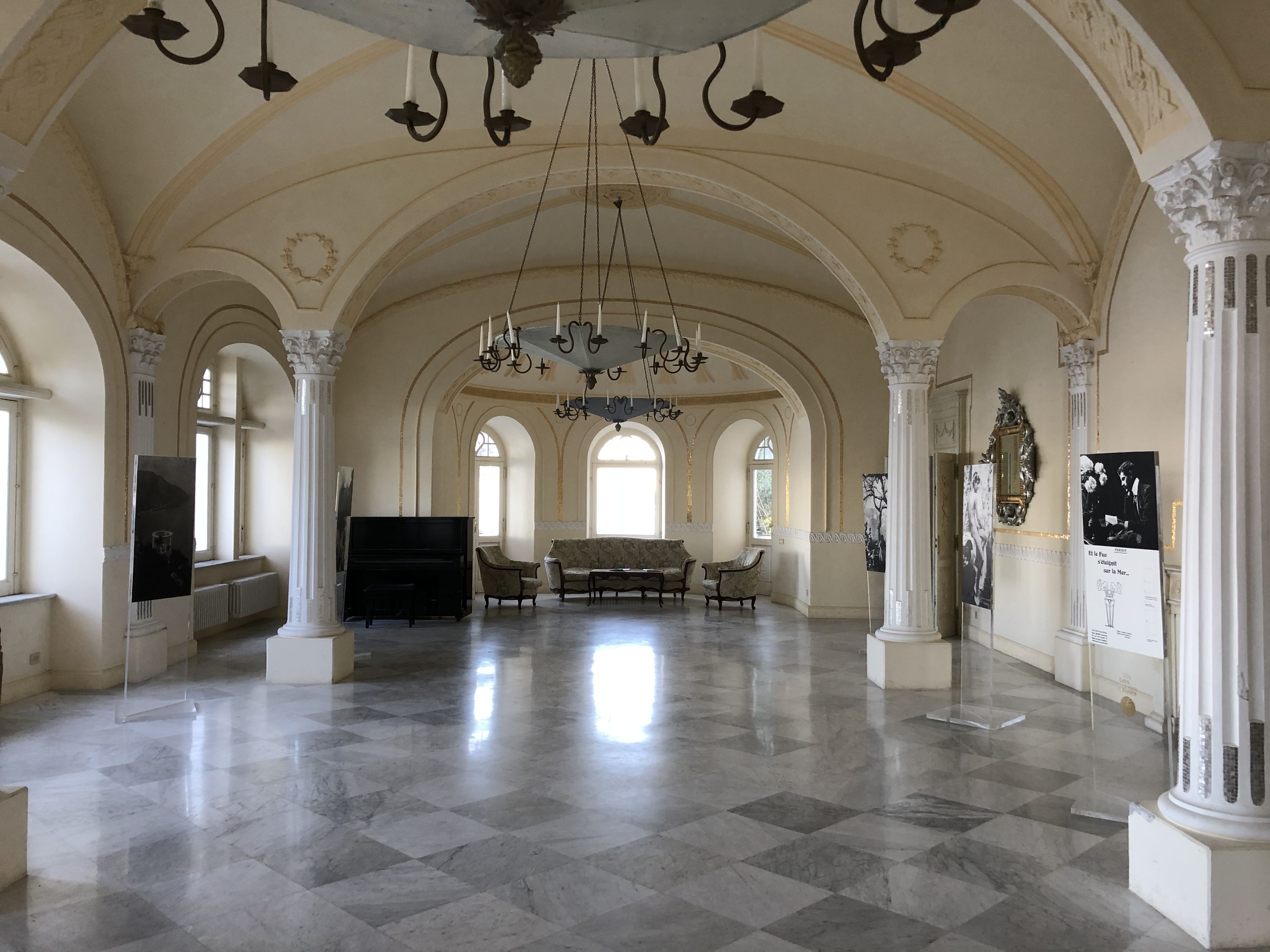 Ornate interior hall of Villa Lysis with vaulted ceilings, marble floor, white columns, chandeliers, and period furniture.
