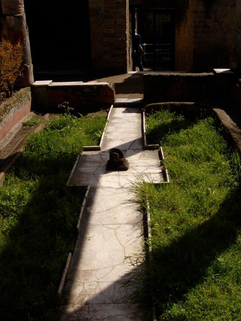 Small peristyle garden in Herculaneum with a central path and grassy beds, surrounded by frescoed walls and low stone borders.