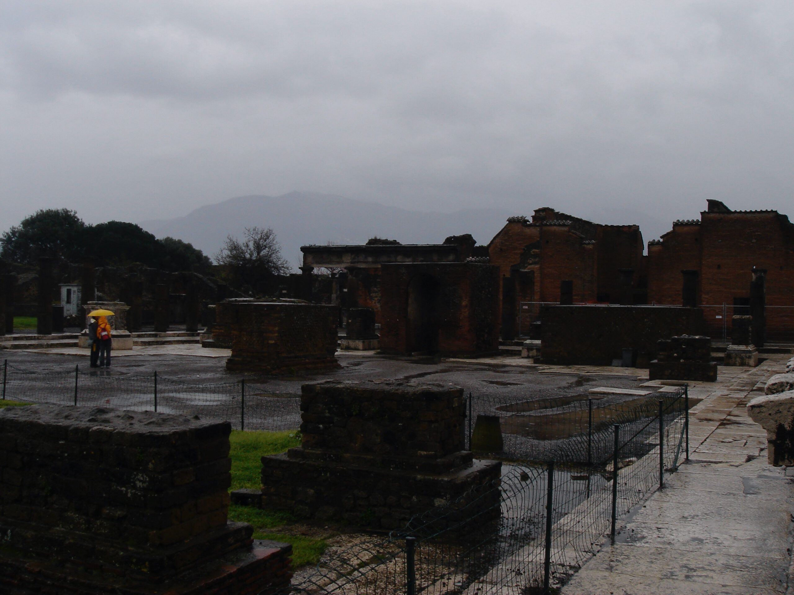 Tourists with umbrellas walk through the rain-soaked ruins of the Forum in Pompeii, with Mount Vesuvius shrouded in mist in the background.